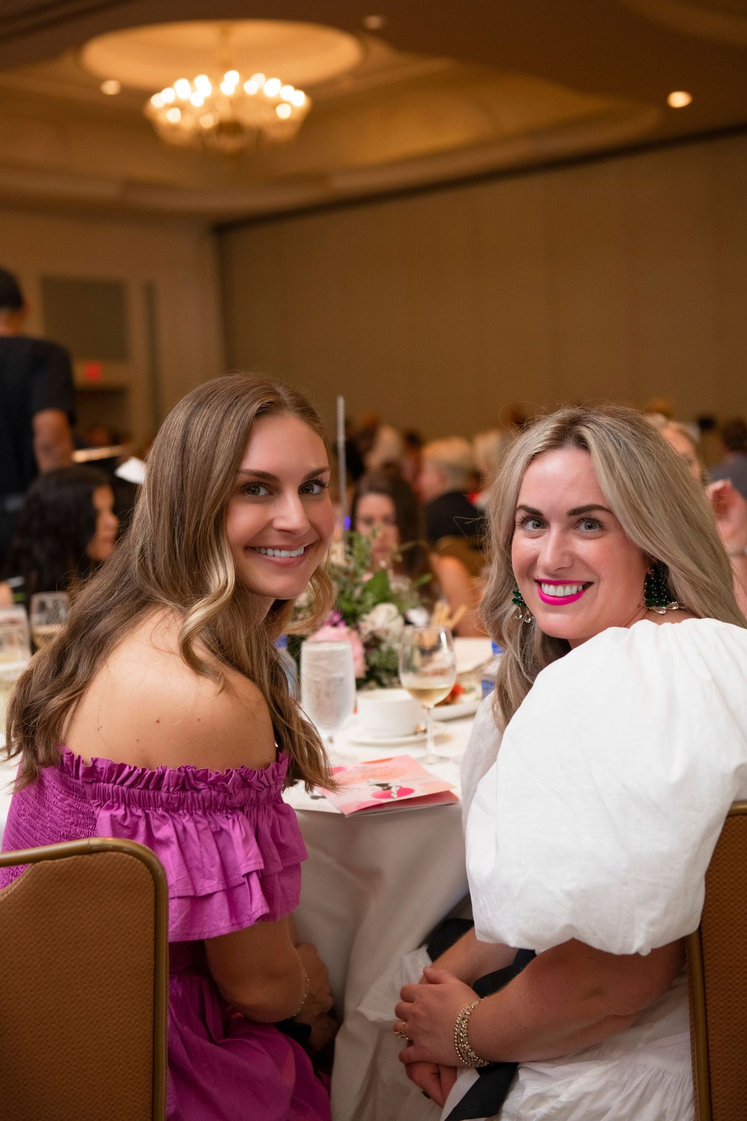 Two smiling women in elegant attire pose at a banquet table during a formal event in a chandelier-lit ballroom.