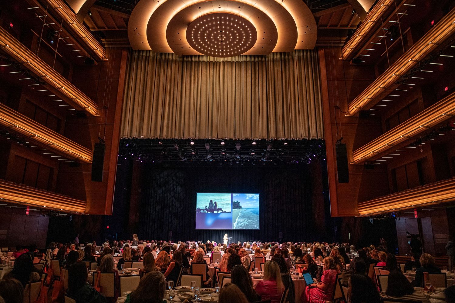 Elegant banquet hall with round tables, guests in formal attire, large stage screen, and ornate ceiling at a gala event.