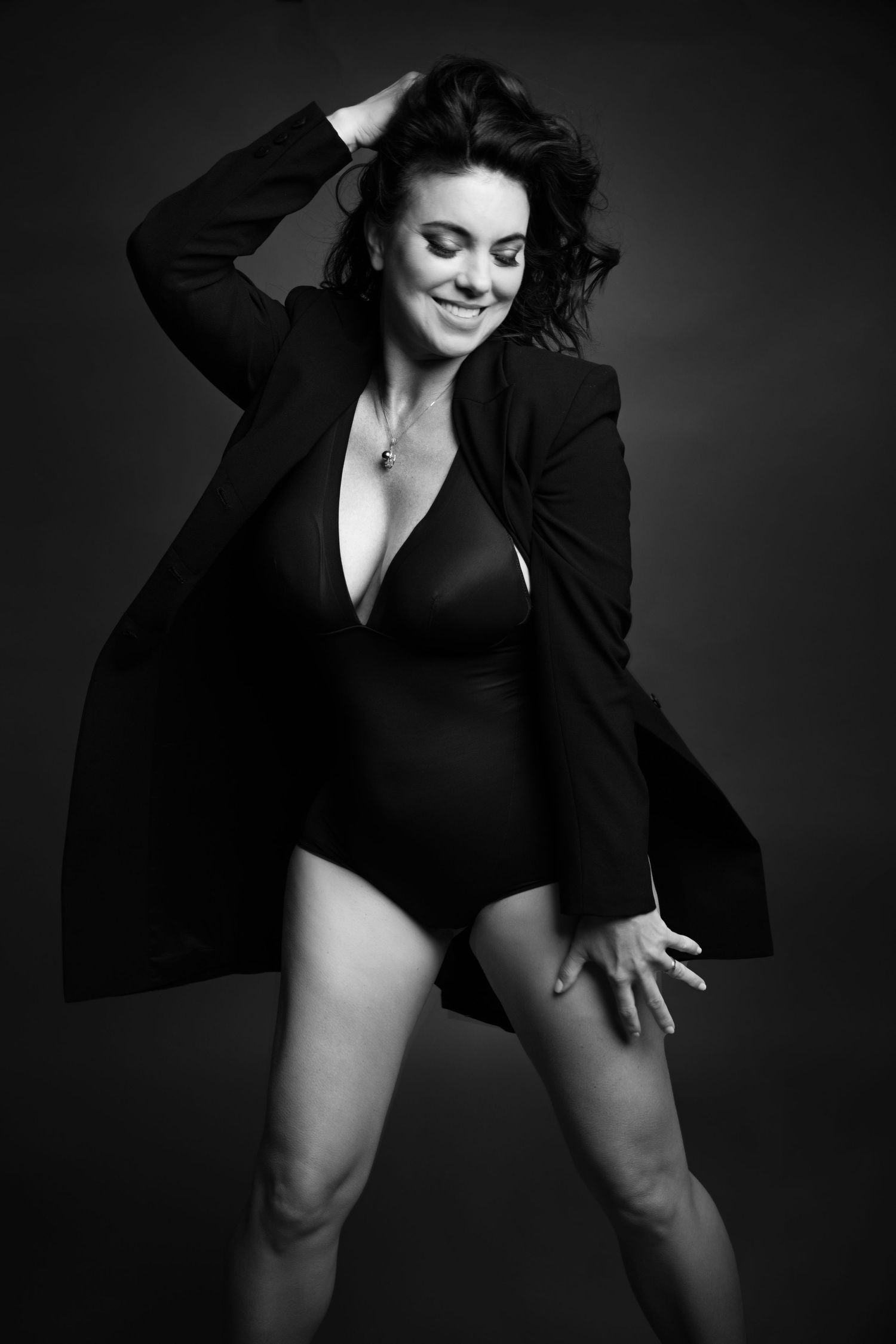 Black and white photo of a smiling woman in a bodysuit and oversized blazer posing confidently against a dark background.