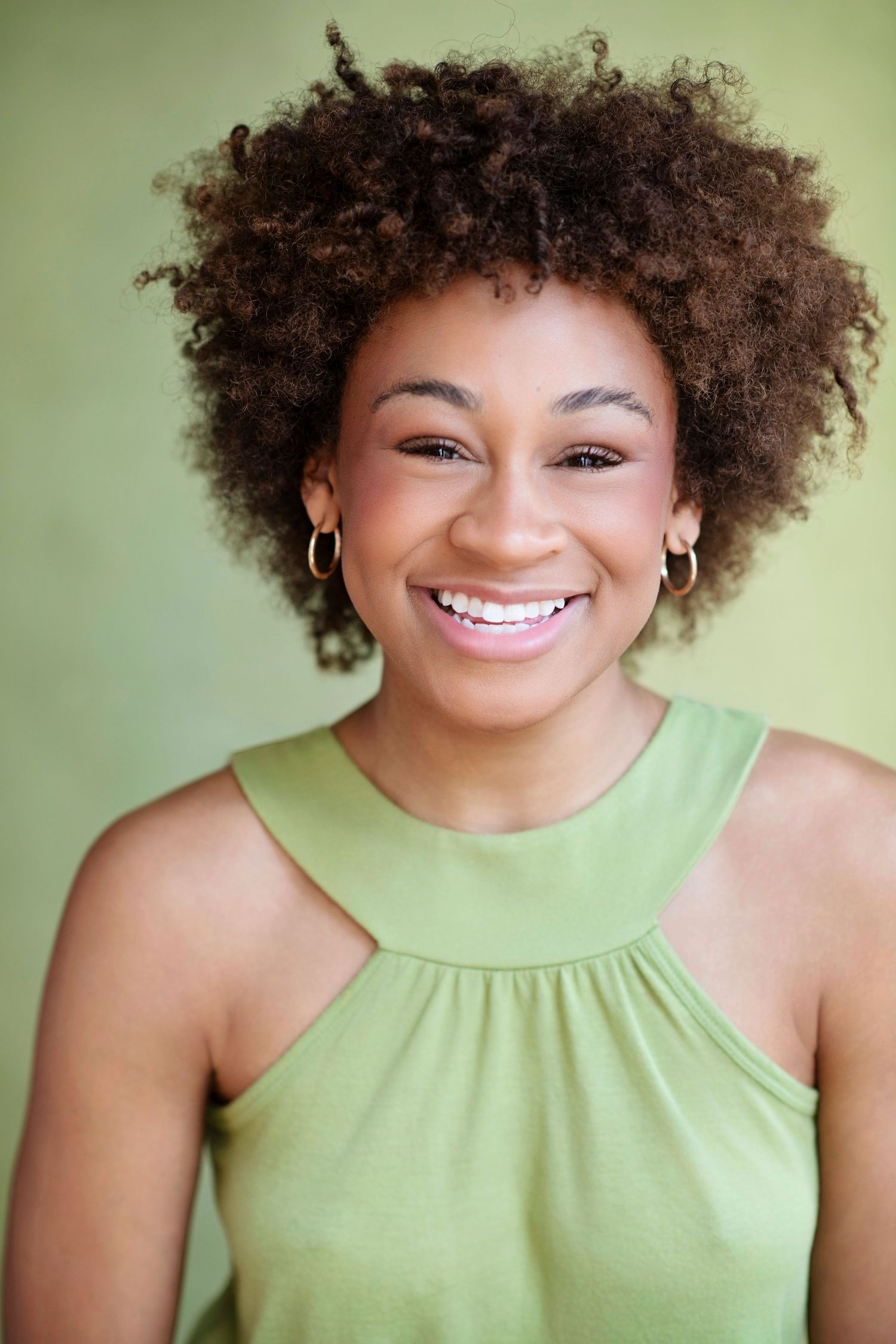 Smiling young Black woman with natural afro hair wearing a green sleeveless top against a green background.