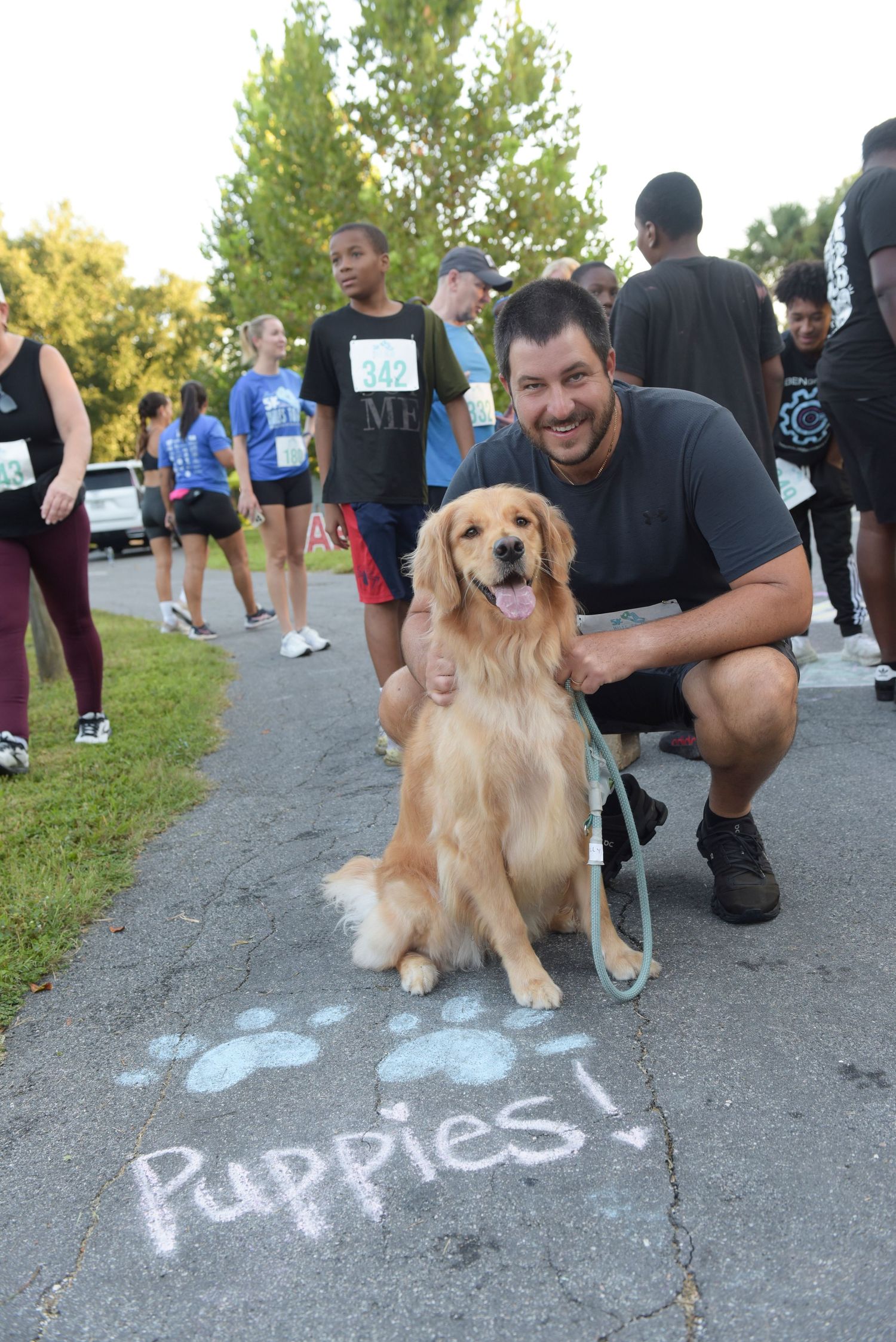 Man crouching with golden retriever dog at outdoor charity run event, paw prints and Puppies text on pavement.