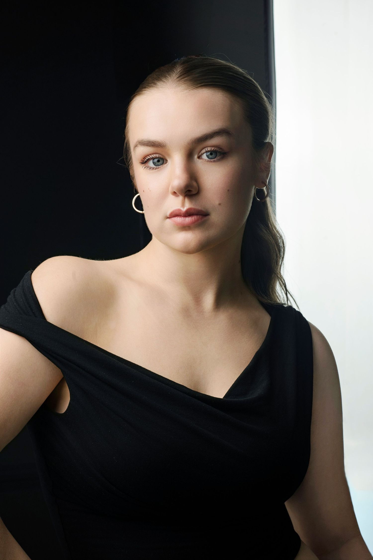 Young woman in off-shoulder black dress with hoop earrings posing against black and white backdrop.