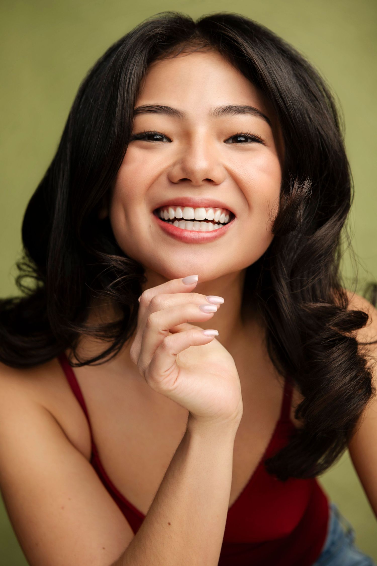 Smiling young Asian woman with wavy black hair wearing a red top against a green background.