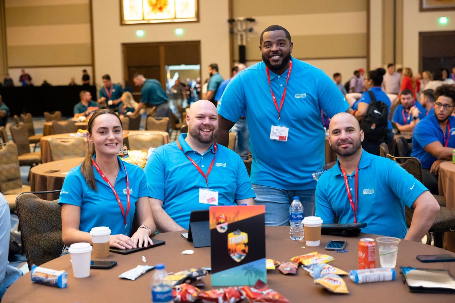 Four smiling professionals in matching blue polo shirts with lanyards gathered around a table at a corporate conference event.