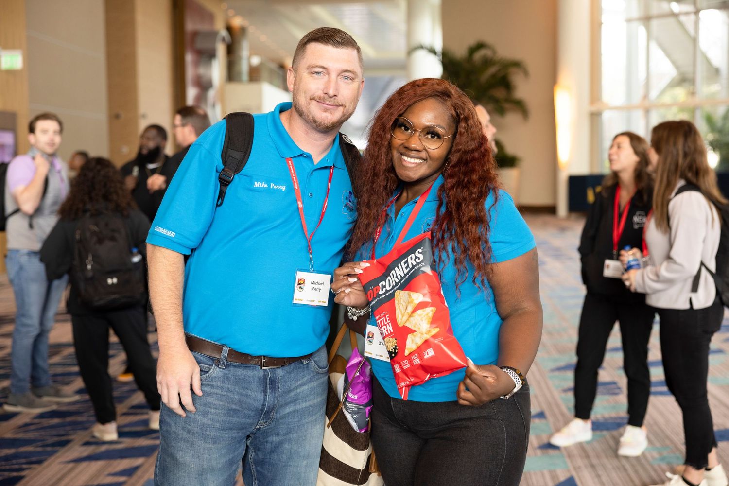 Two colleagues in blue shirts smile together at a conference, one holding a snack bag, with attendees in the background.