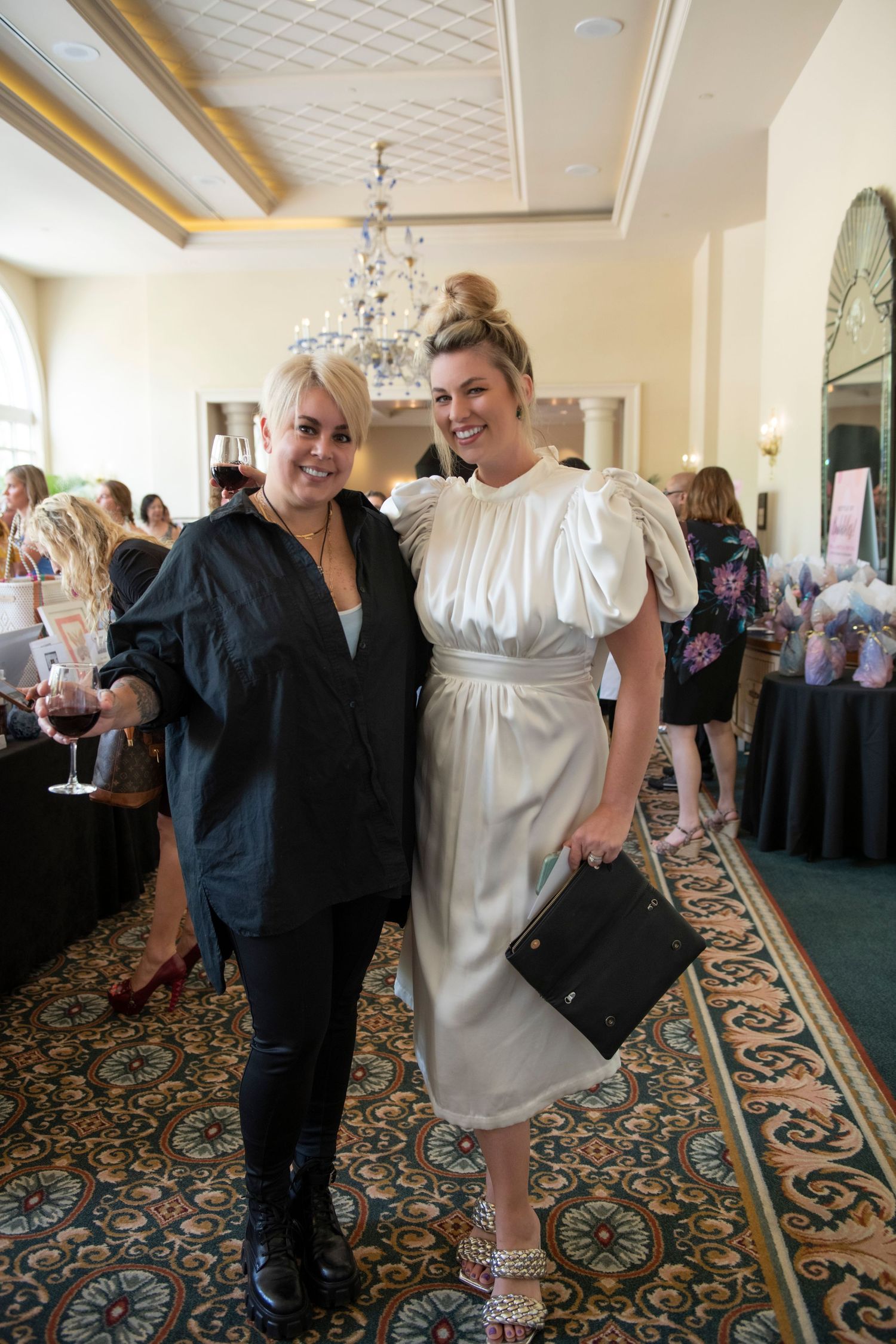 Two stylish women posing together at upscale event, one in black outfit and one in white puff-sleeve dress.