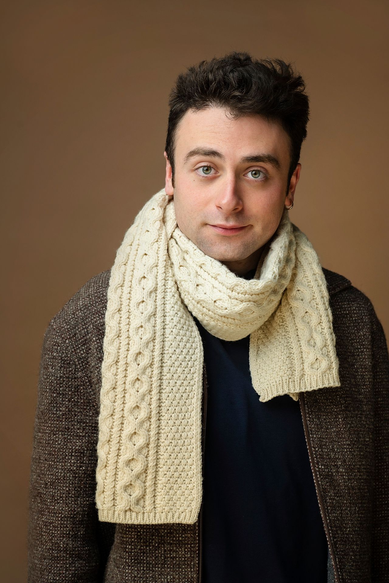 Handsome man wearing a cream knit scarf and brown jacket poses for a warm-toned studio portrait.