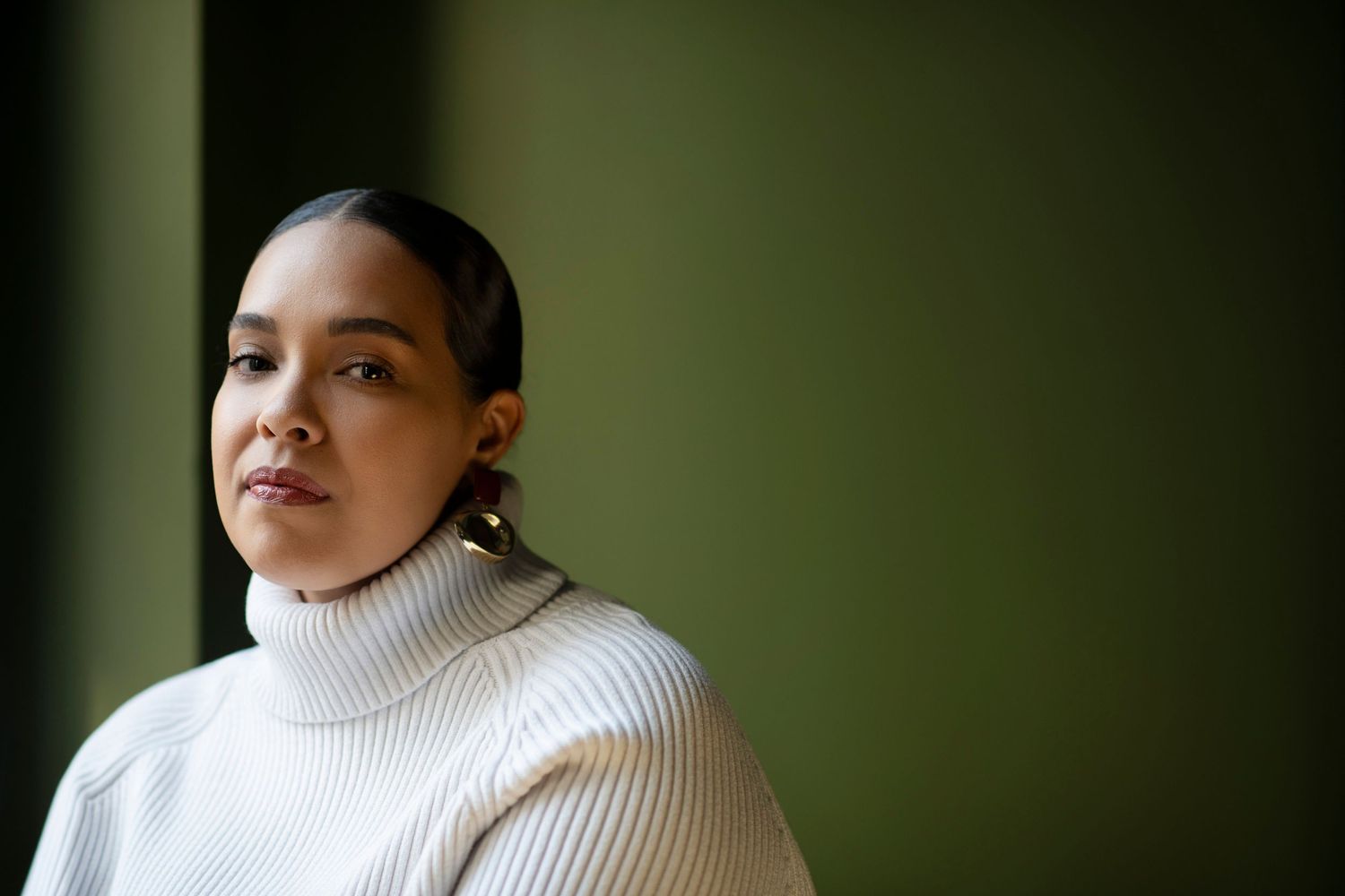 Young woman in white turtleneck sweater gazing thoughtfully against a dark green background.