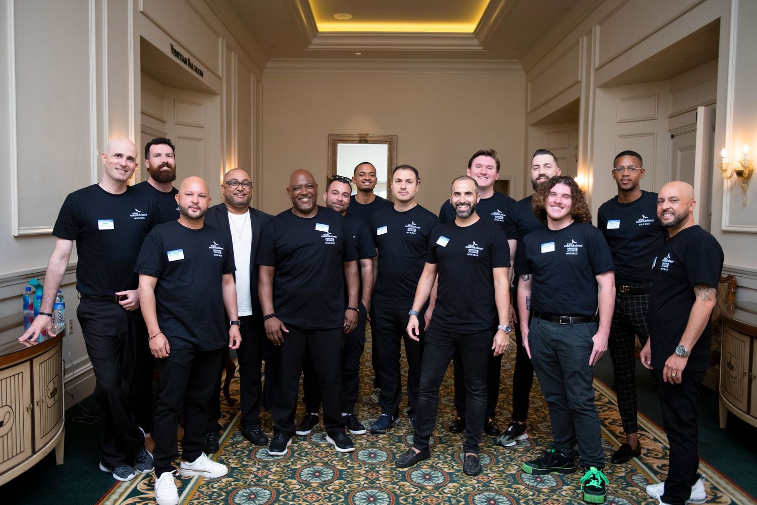 Group of men in matching black t-shirts posing together in an elegant hallway at a formal event.