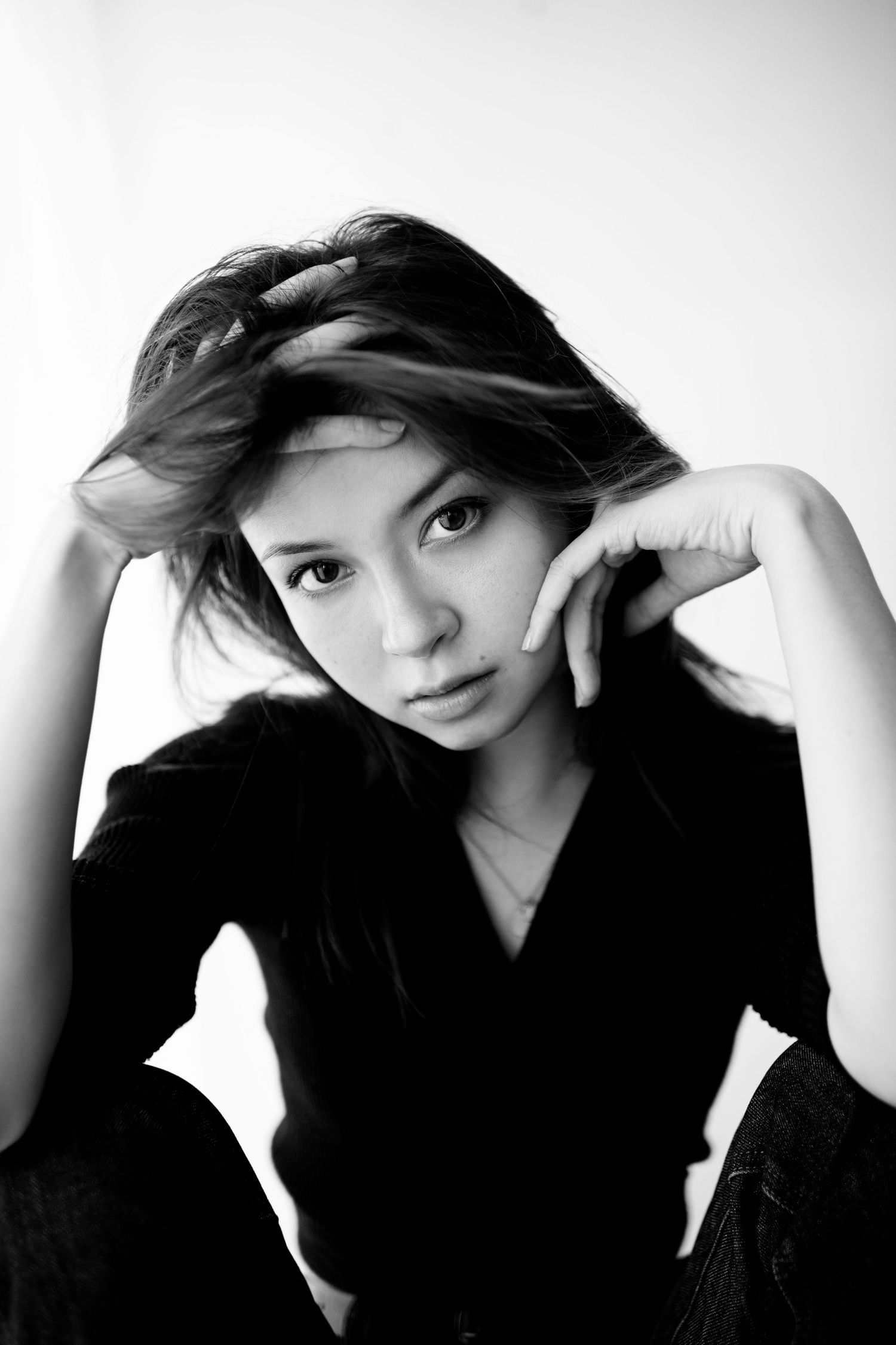 Black and white portrait of a young Asian woman in a black outfit, seated with hand near face, hair flowing.