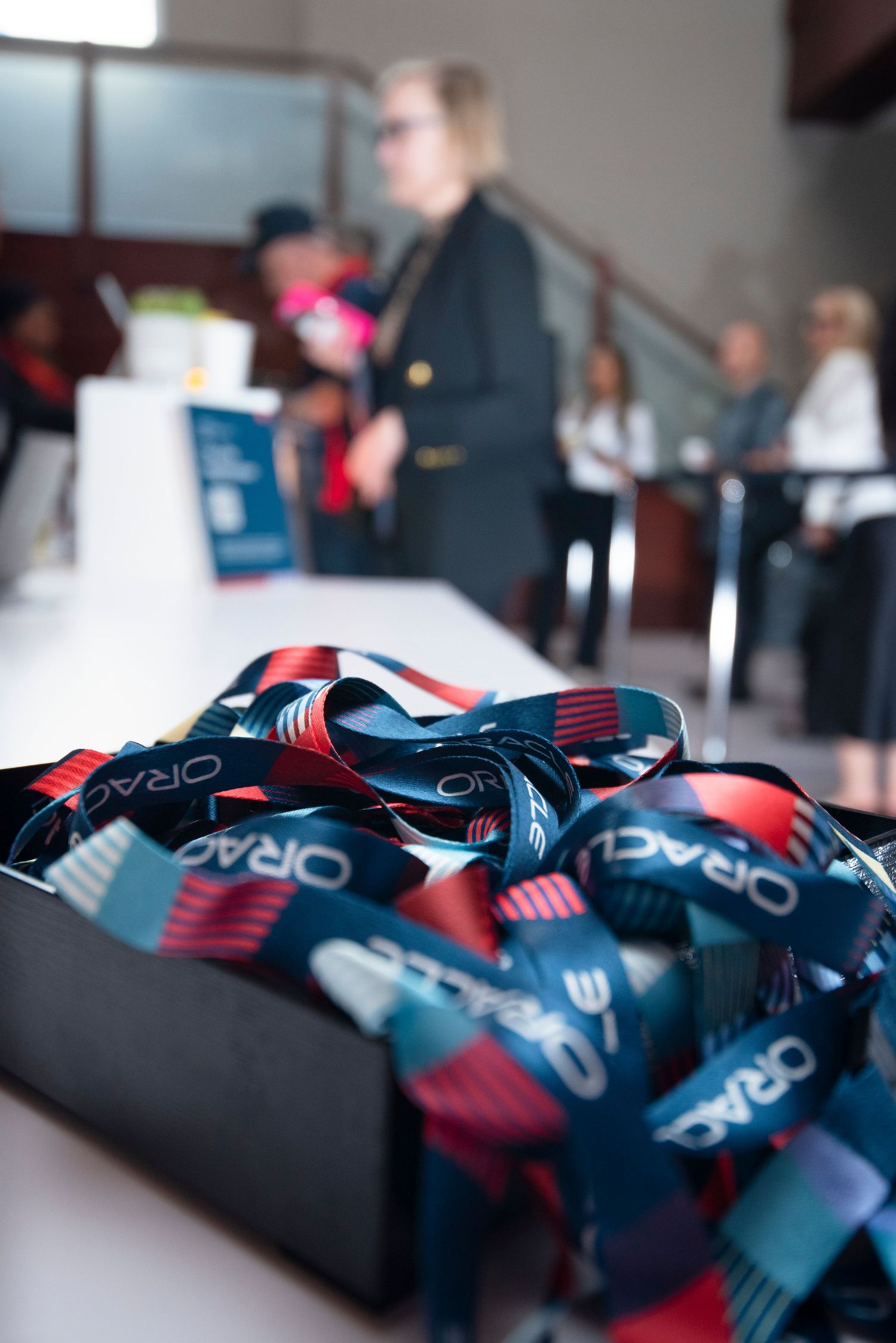 A box of Oracle branded lanyards in red and navy blue at a corporate registration desk with attendees in background.