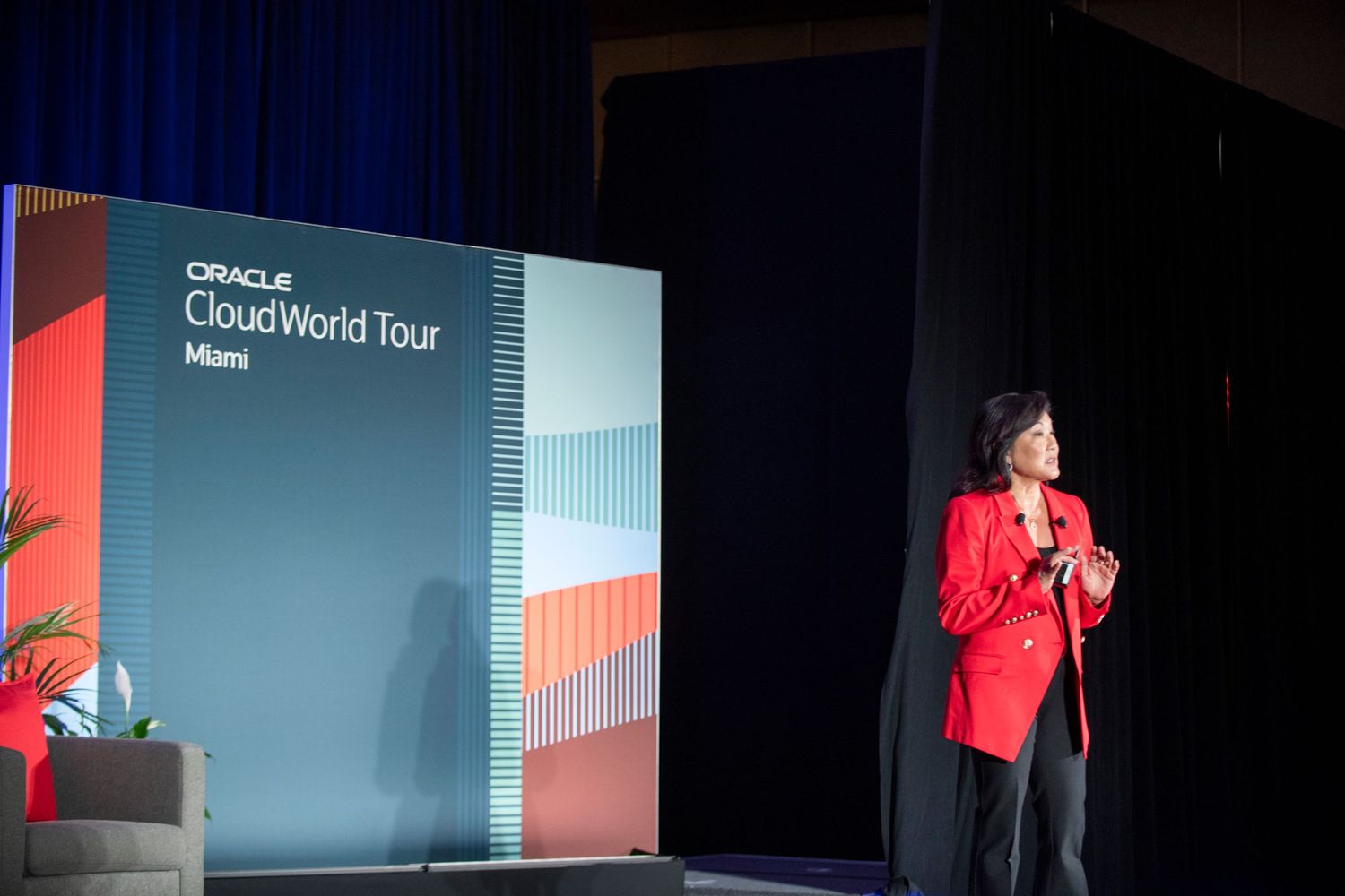 Speaker in red blazer presents at Oracle CloudWorld Tour Miami event on a dark stage with branded backdrop.