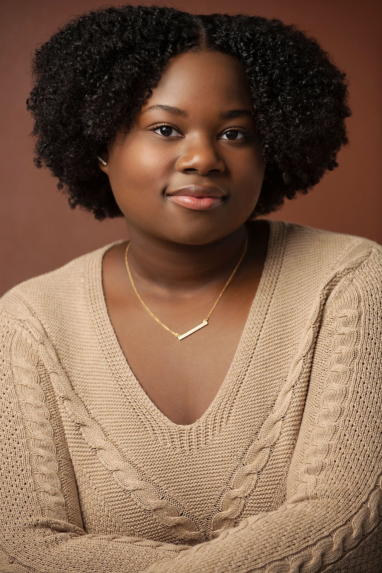 Young Black woman with natural hair wearing a beige cable-knit sweater and gold necklace.