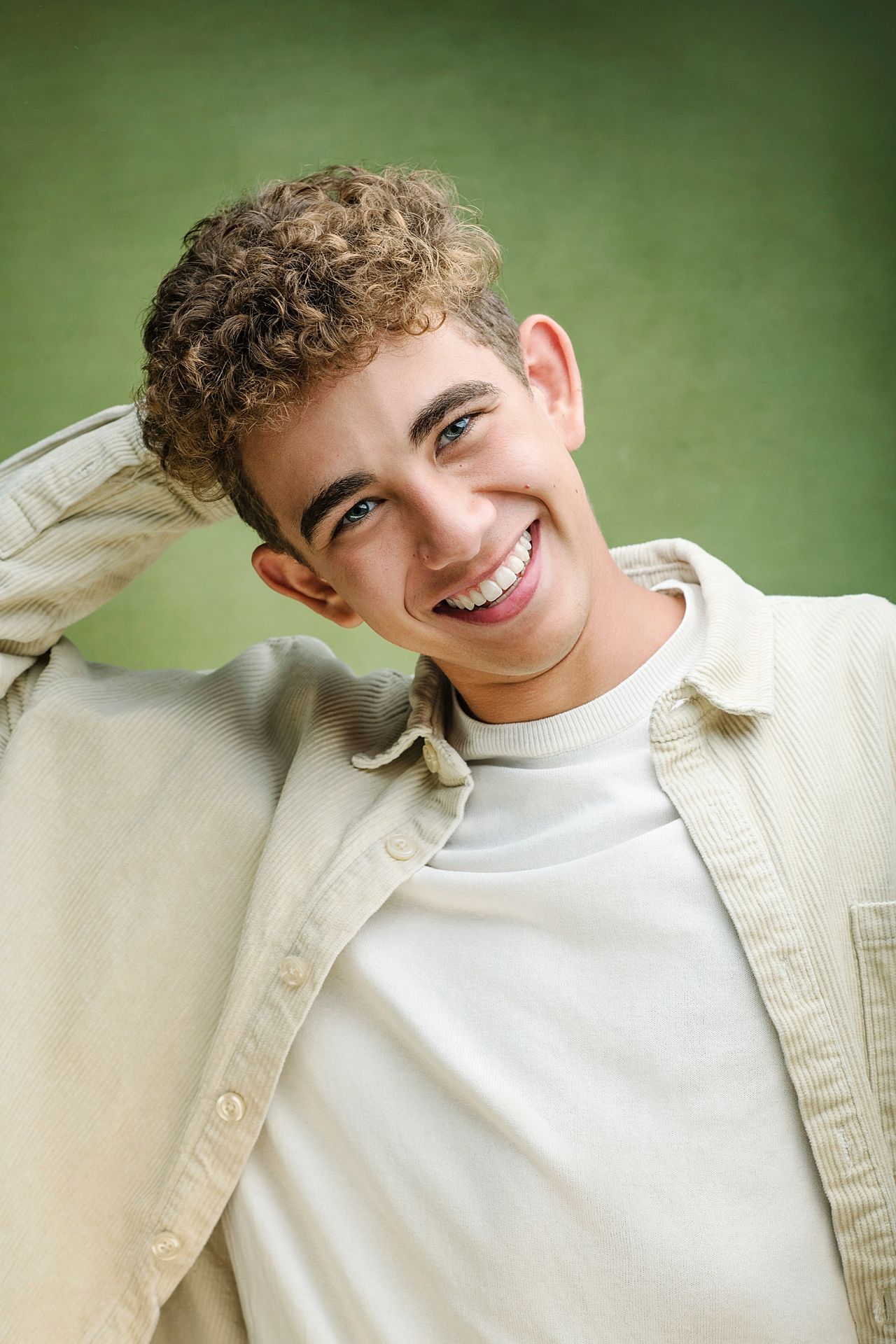 Smiling young man with curly hair wearing beige shirt poses against green background.