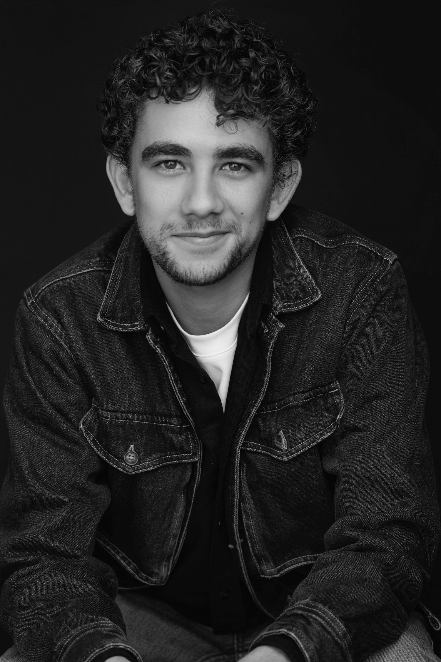 Smiling young man with curly hair wearing a denim jacket in a black and white portrait photo.