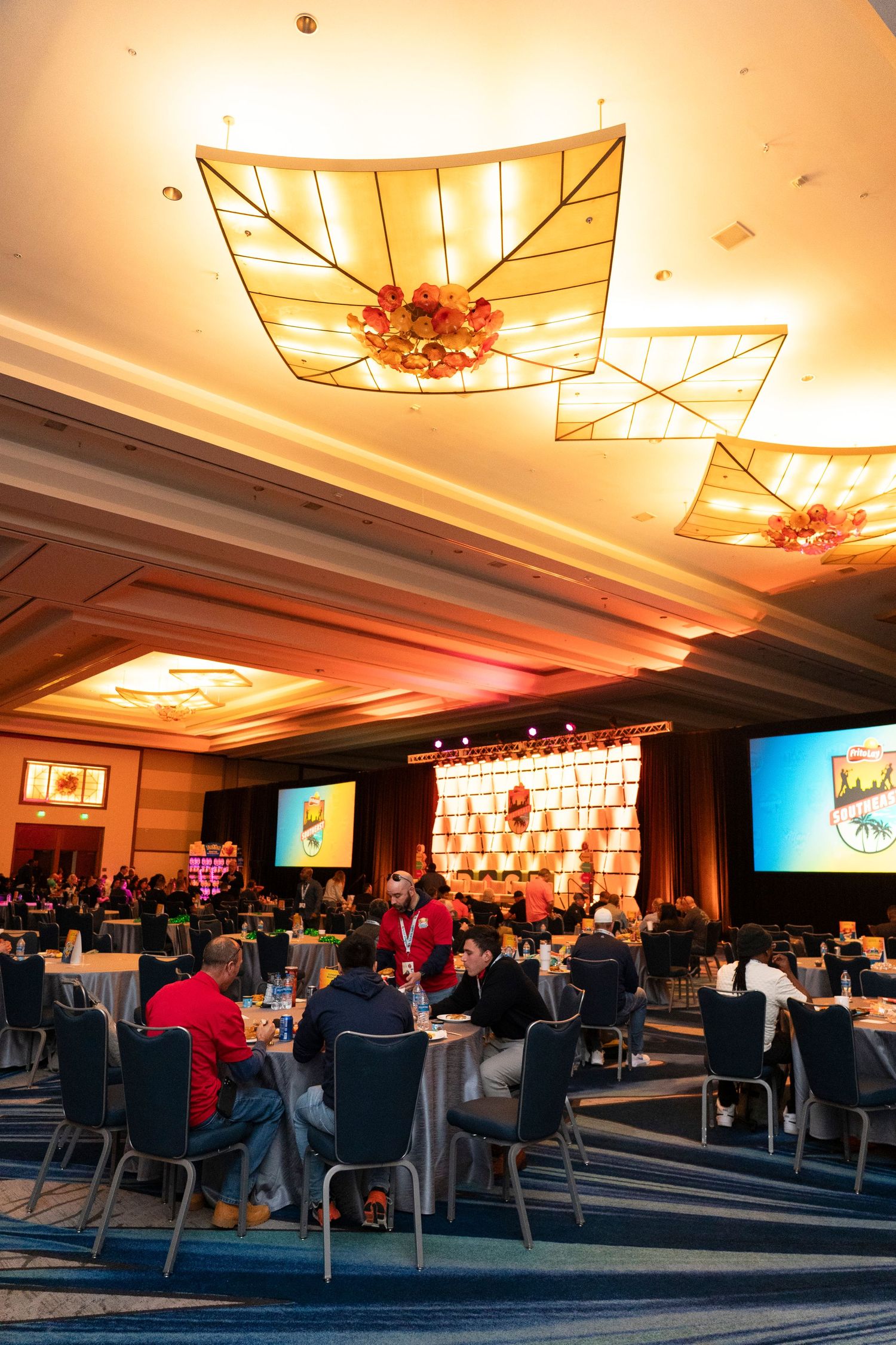 Elegant hotel ballroom event with round tables, attendees networking, colorful screens, and decorative ceiling lights.