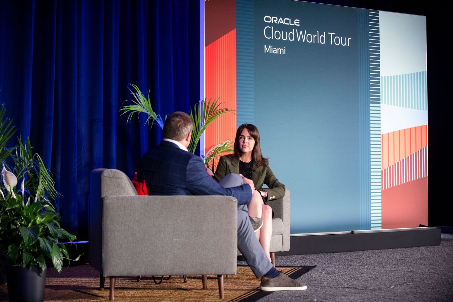 Two professionals engage in a panel discussion at Oracle CloudWorld Tour Miami event on a blue-lit stage.