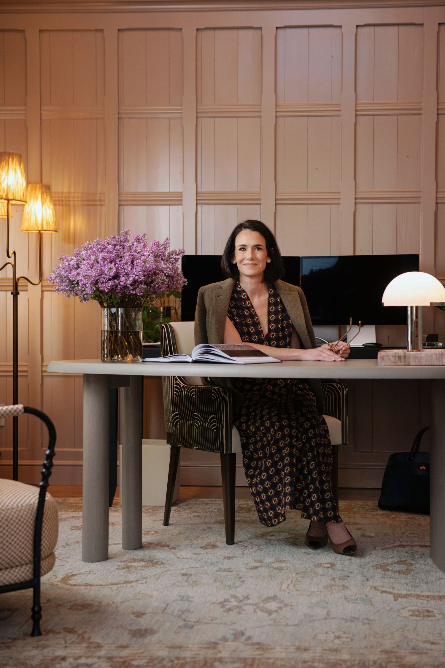 Professional woman in patterned dress sits at elegant desk with purple flowers in a warm wood-paneled office.