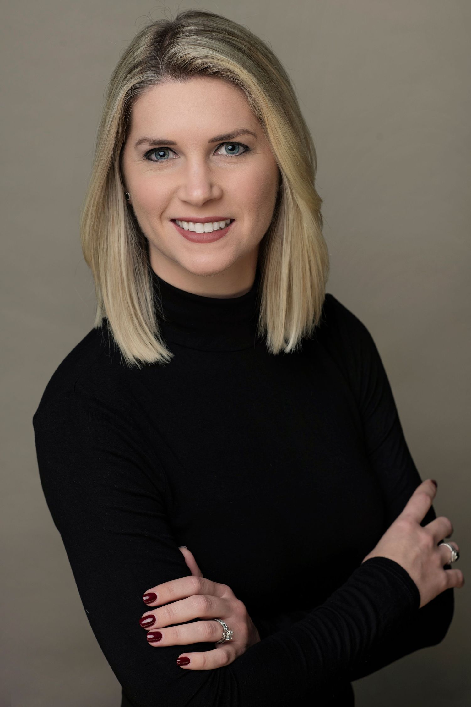 Professional headshot of a smiling blonde woman in a black turtleneck with arms crossed against a gray background.