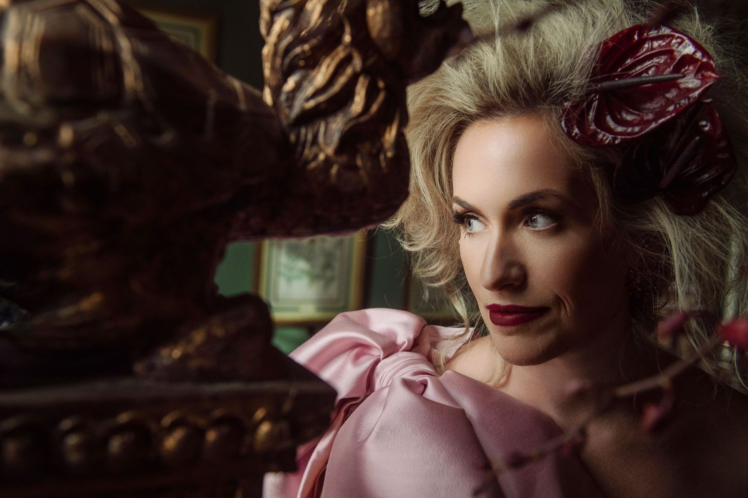 Glamorous blonde woman in pink ruffled gown with decorative hair accessory gazes sideways in ornate setting.