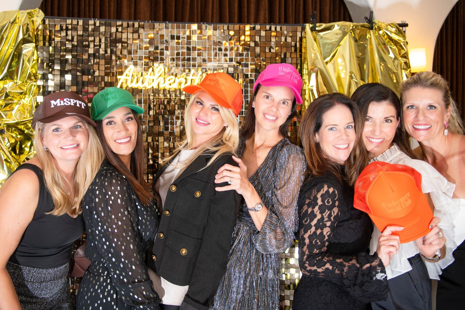 Seven women smiling together at a party, several wearing colorful baseball caps, posing in front of a gold backdrop.