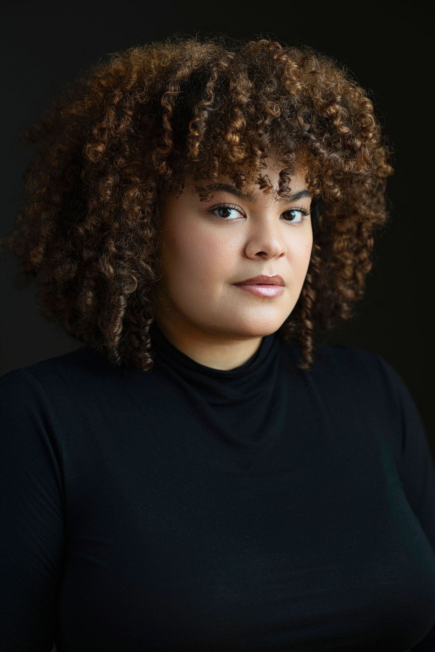 Young woman with curly brown hair wearing a black turtleneck poses against a dark background.