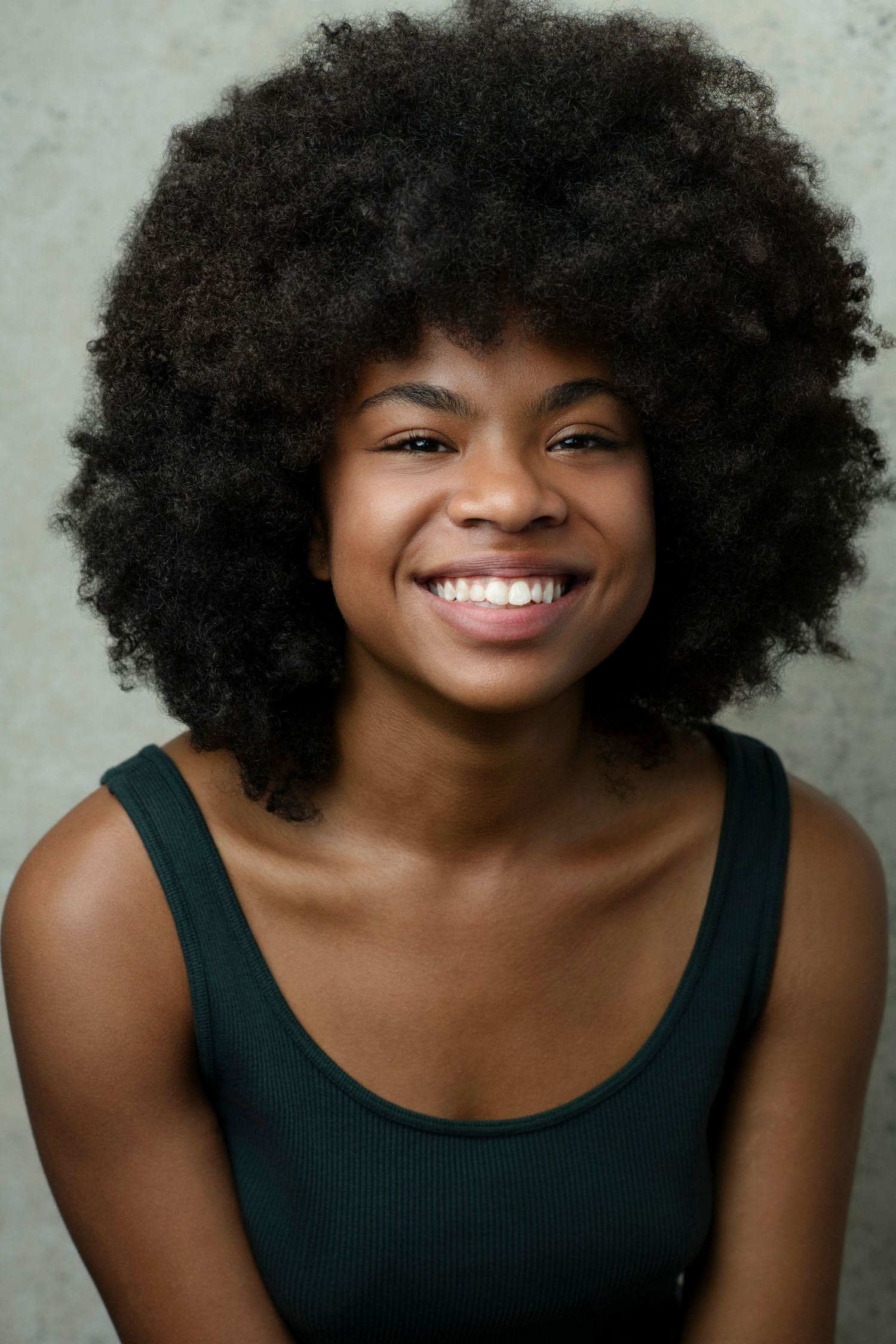 Young Black woman with natural afro hair smiling broadly, wearing a dark green tank top against a gray background.