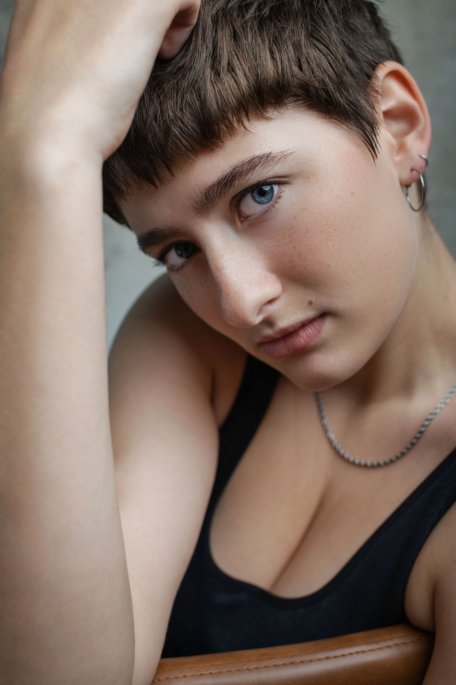 Young person with short brown hair, blue eyes, hoop earrings, and silver necklace posing in a black tank top.