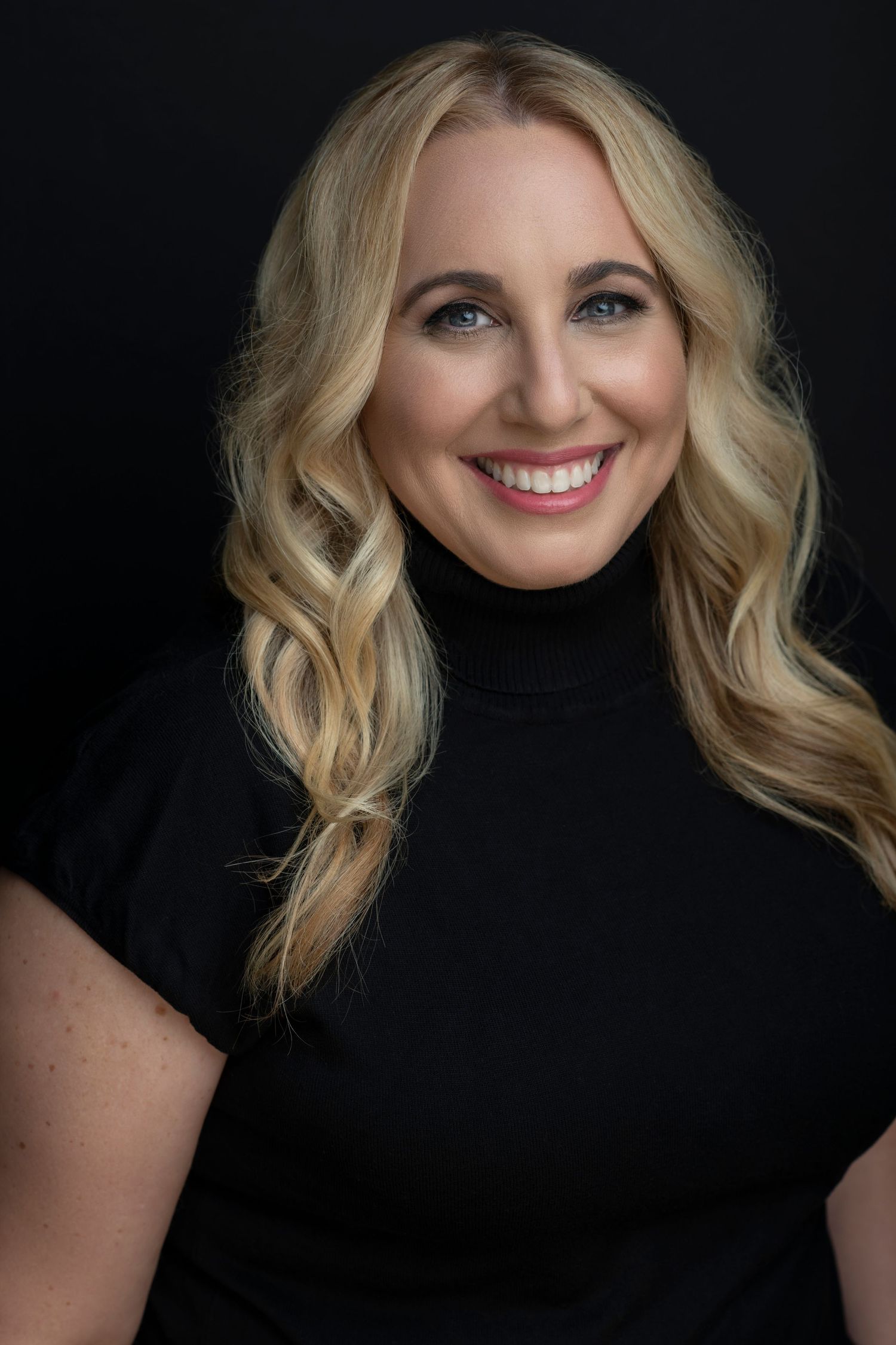Smiling blonde woman with wavy hair wearing a black turtleneck top, professional headshot on dark background.