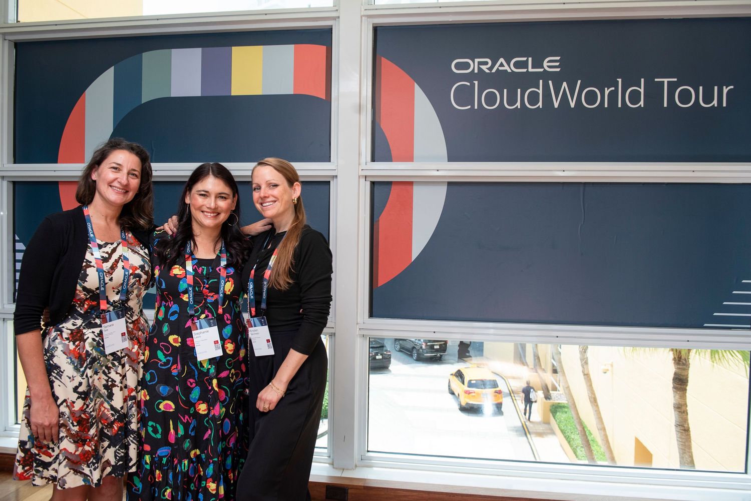 Three women smiling together at Oracle Cloud World Tour event, standing in front of branded signage.