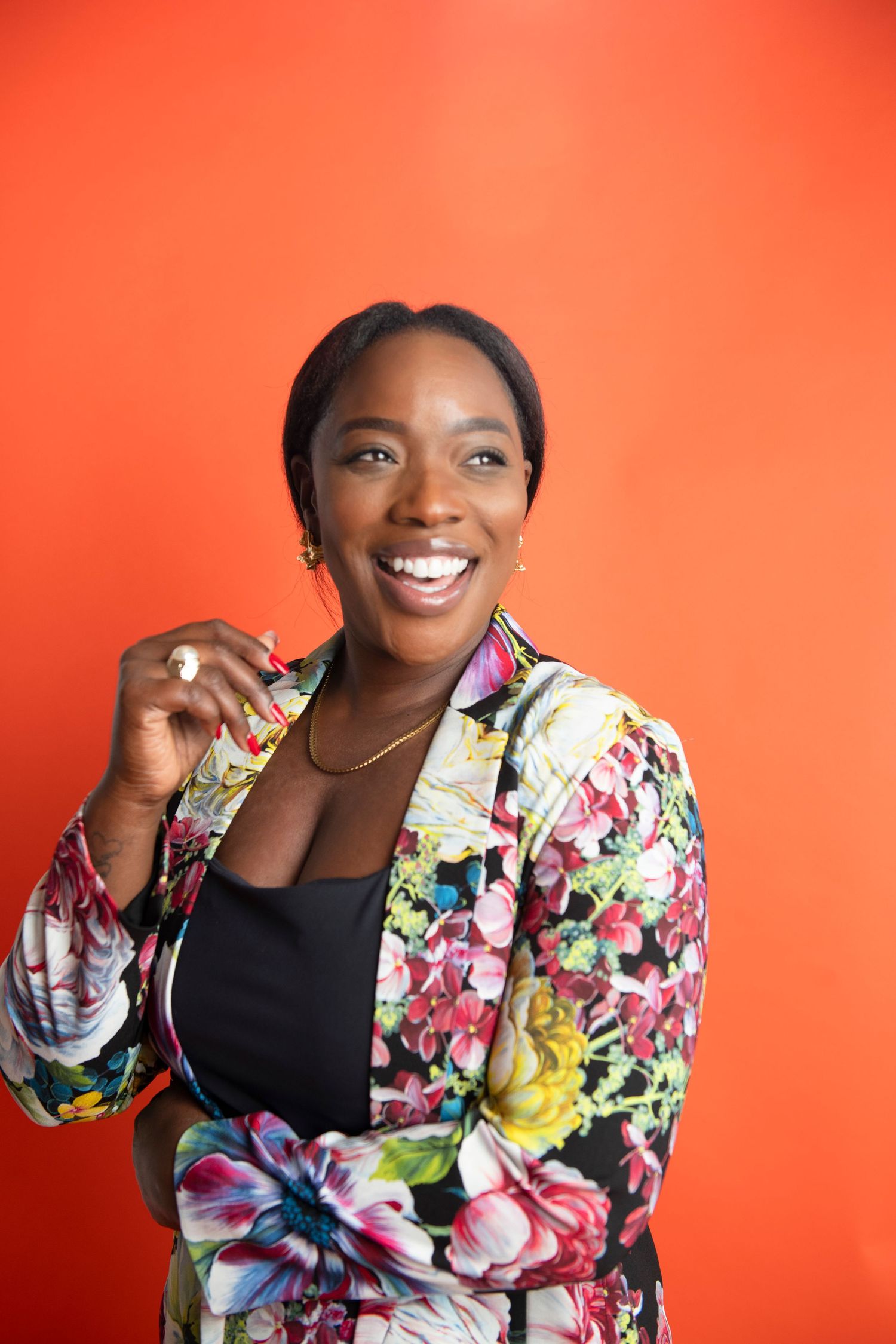 Smiling Black woman in colorful floral blazer over black top poses against vibrant orange background.