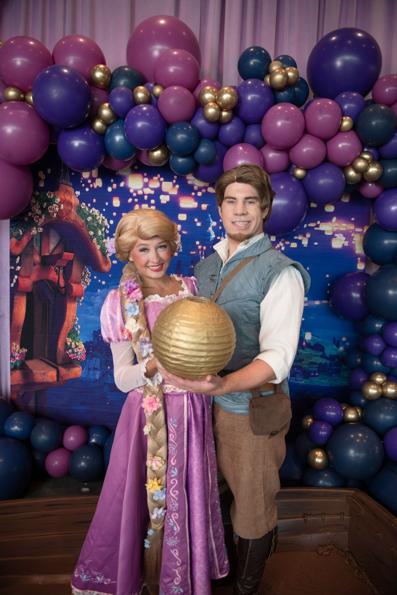 Rapunzel and Flynn Rider cosplay couple holding gold lantern at Tangled themed party with purple balloon arch.