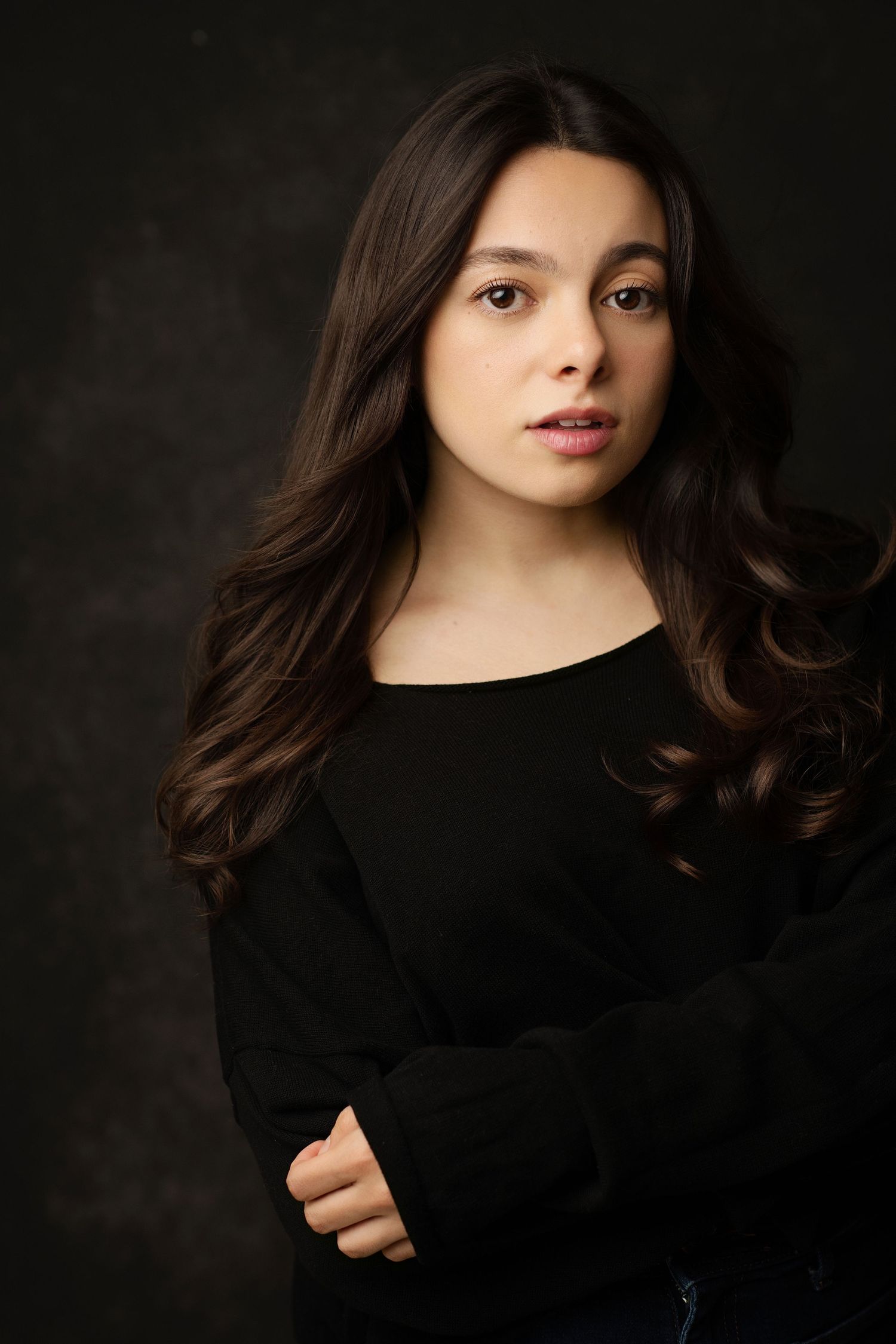 Young woman with long dark wavy hair wearing a black top, posing against a dark studio background.