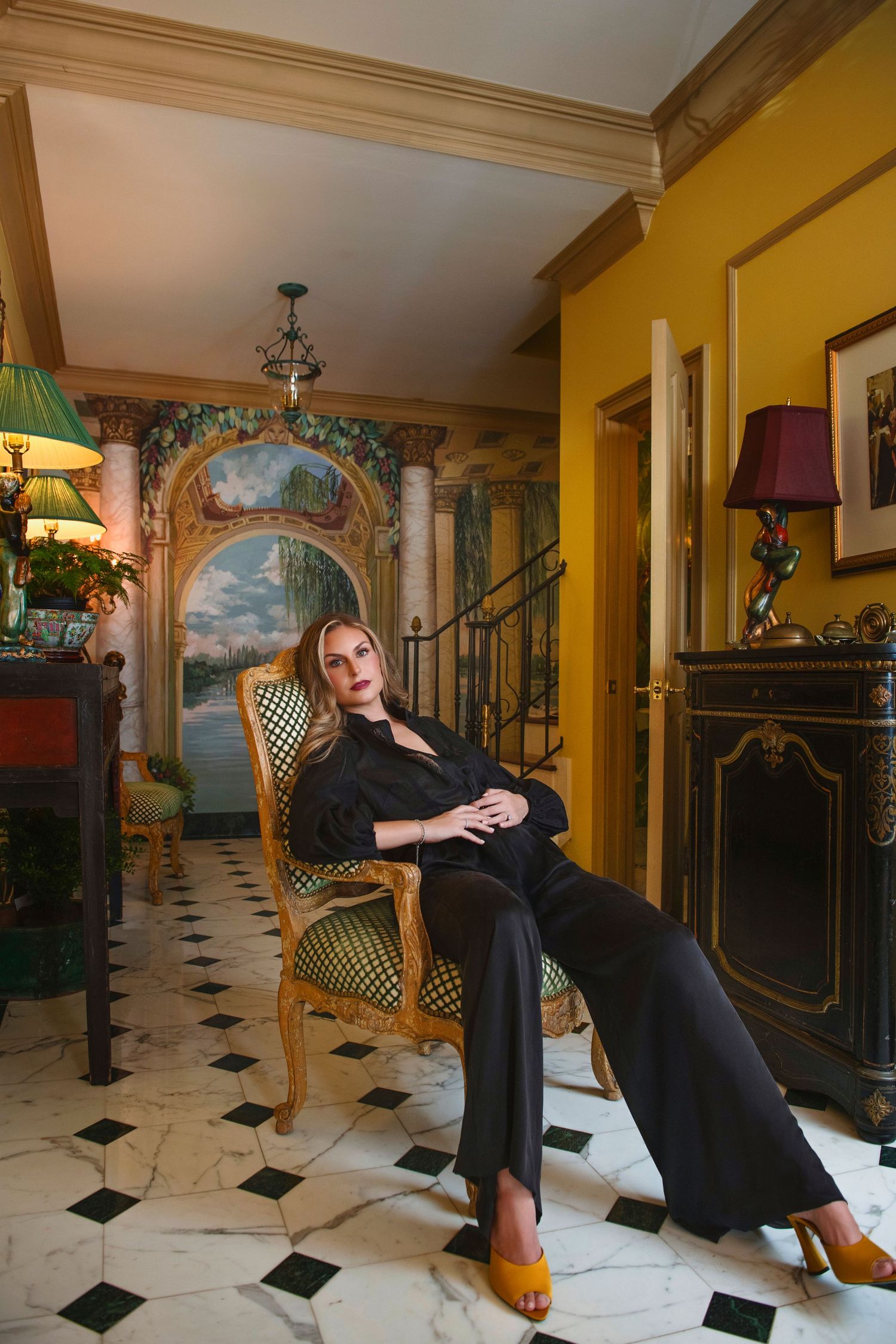 Elegant woman in black pantsuit seated on gilded chair in luxurious yellow classical interior.