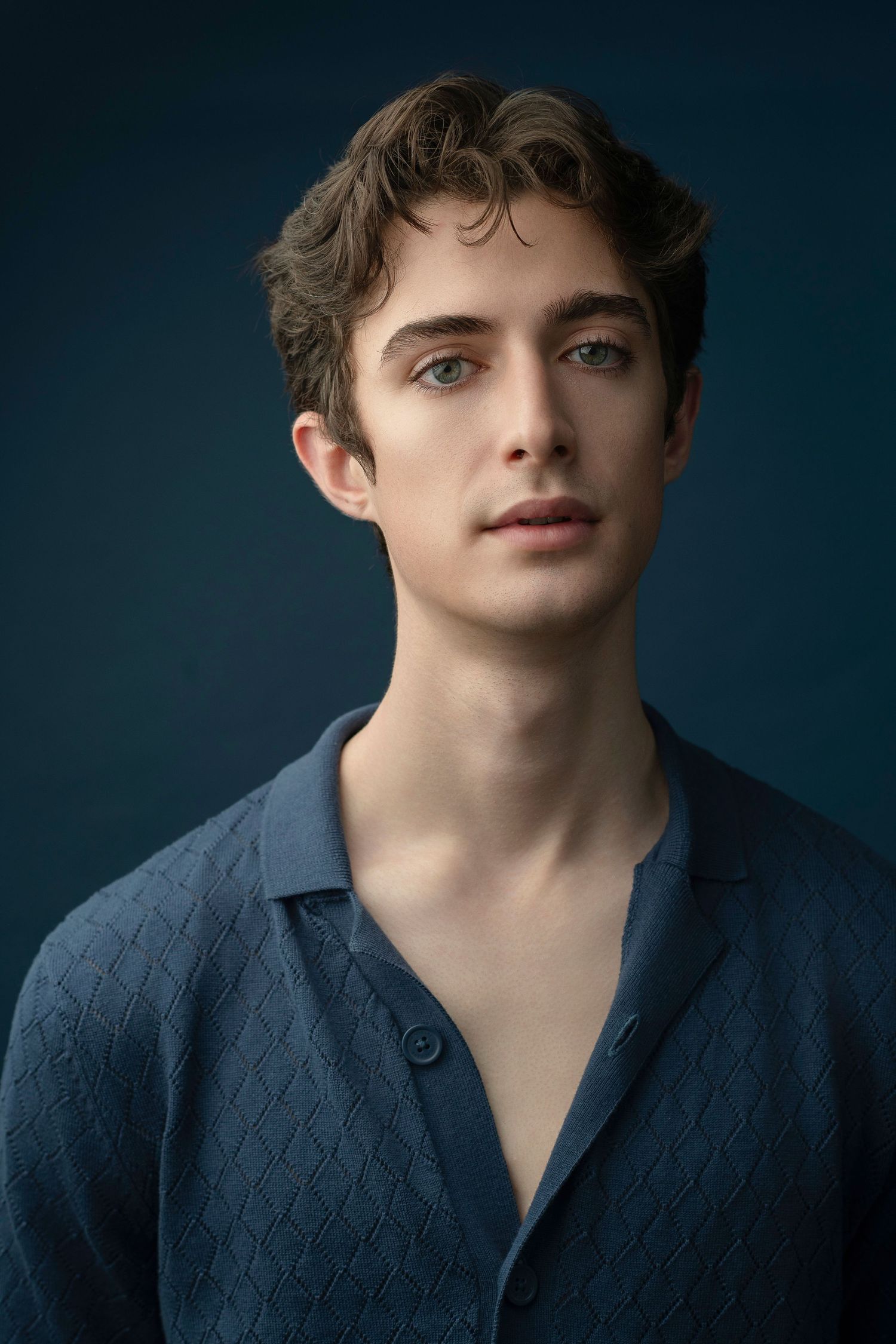 Young man with wavy brown hair wearing a navy blue textured button-up shirt against a dark teal background.