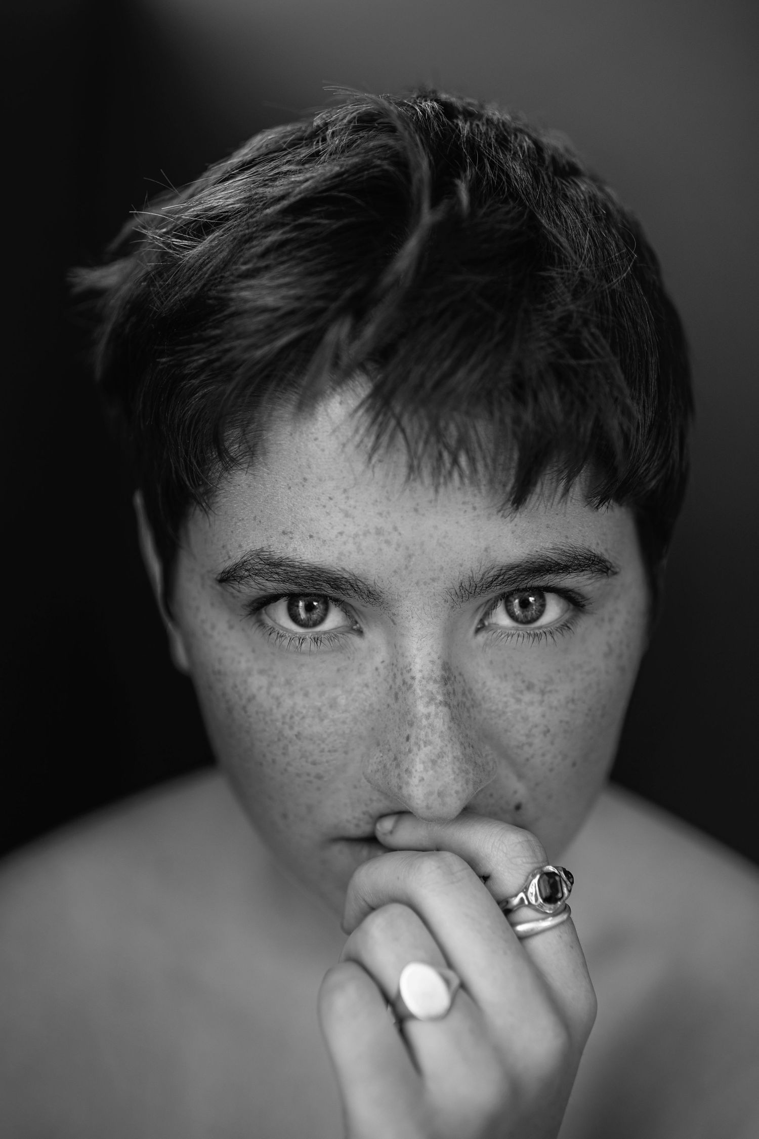 Black and white portrait of freckled person with short dark hair holding ring to lips.