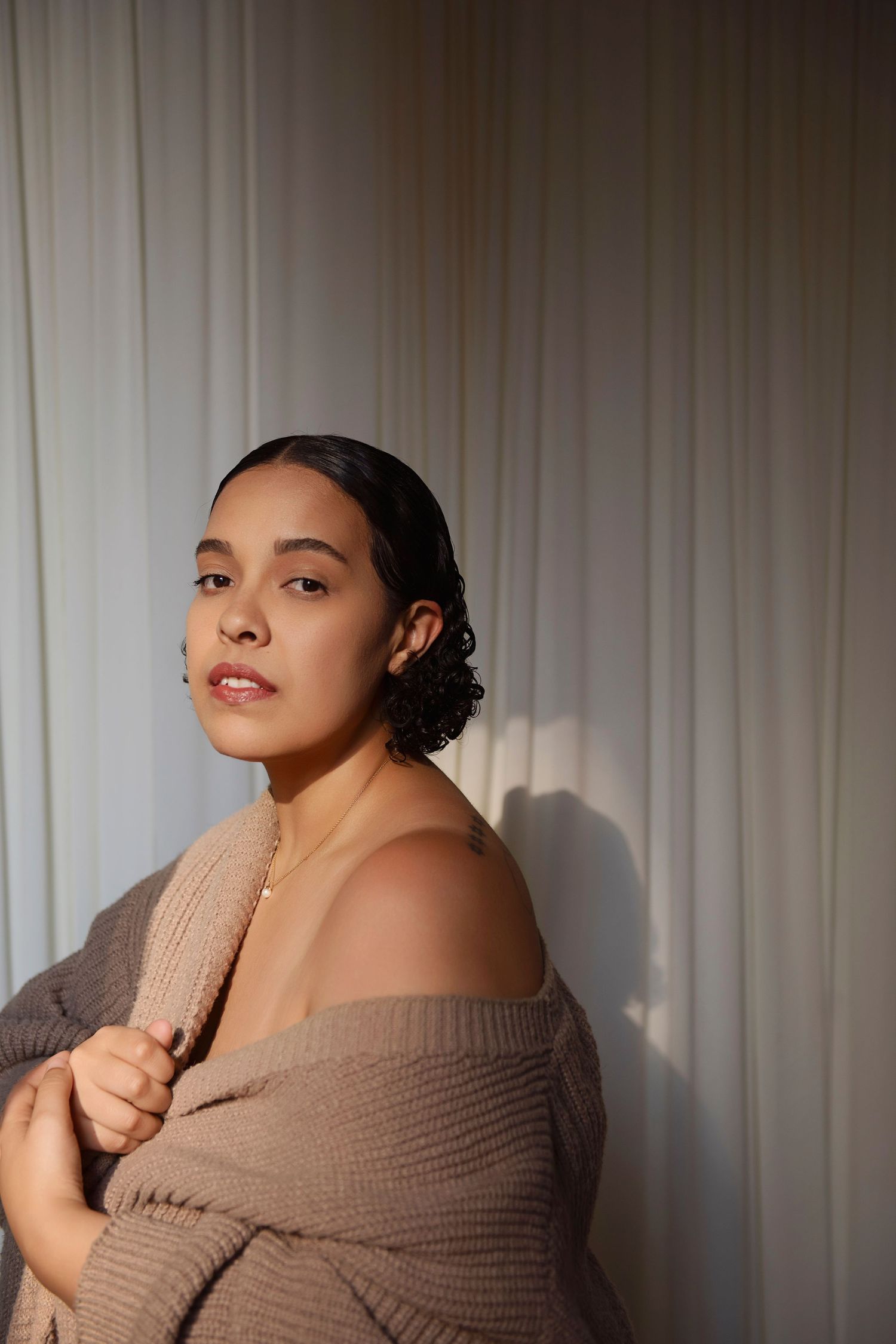 Young woman in off-shoulder beige knit sweater poses gracefully against white curtain backdrop in soft natural light.