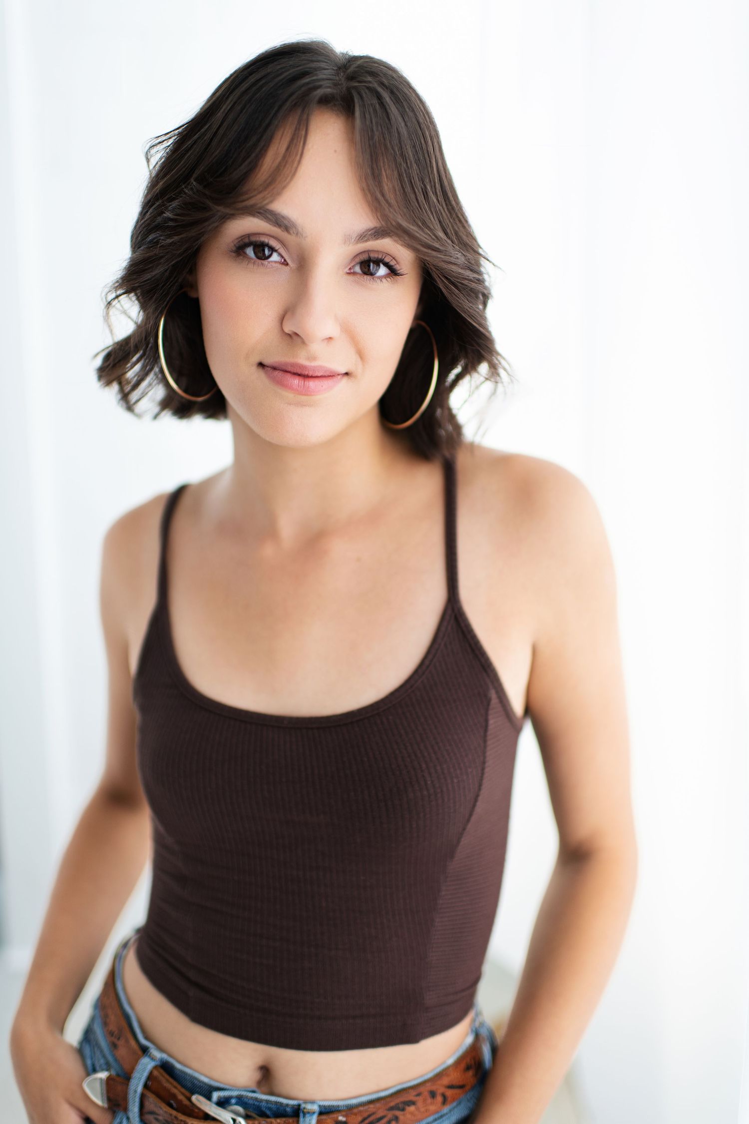 Young woman with short dark hair wearing gold hoop earrings and a brown crop top with jeans, posing against white background.
