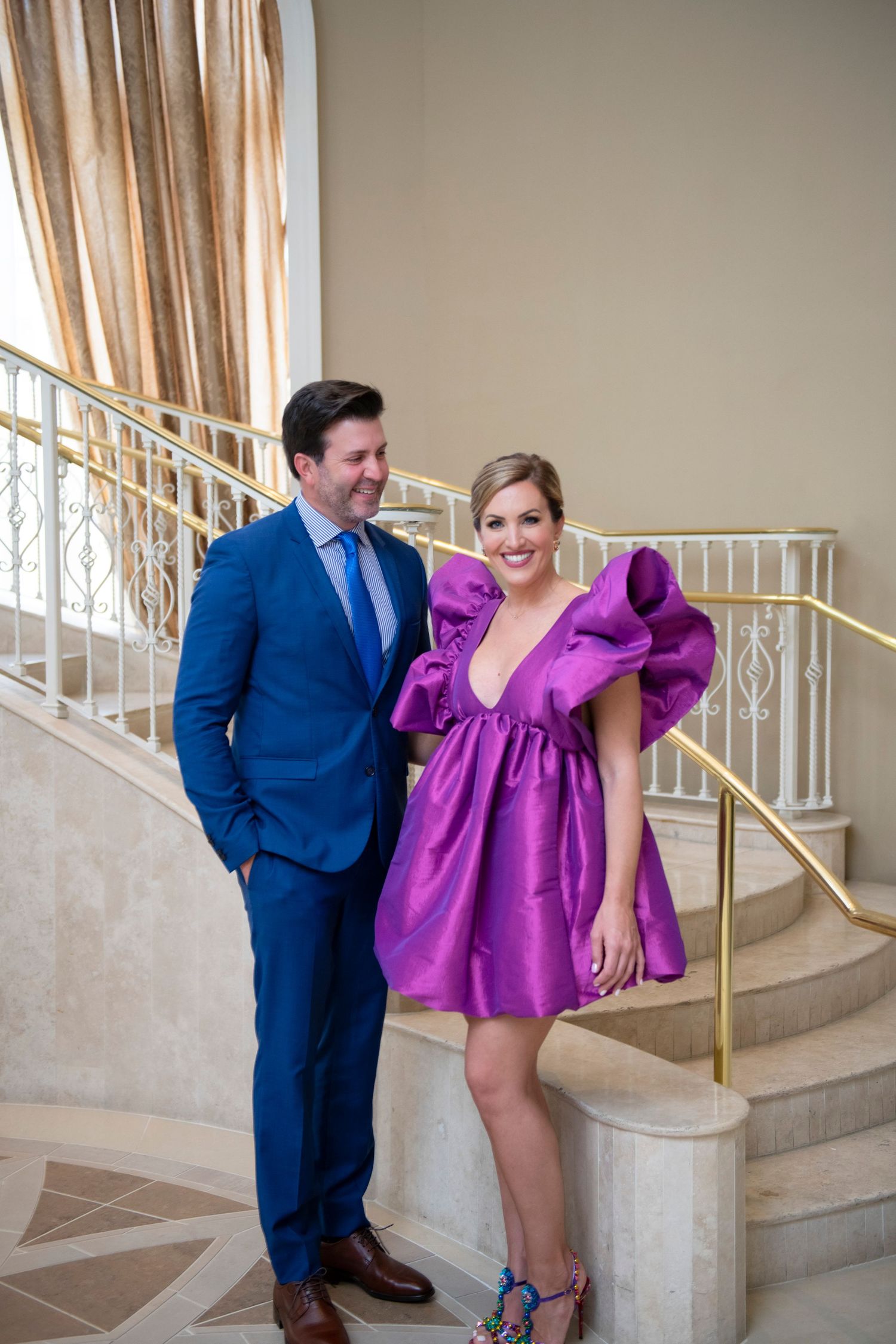A man in a blue suit and woman in a purple mini dress pose together on an elegant staircase with gold railings.