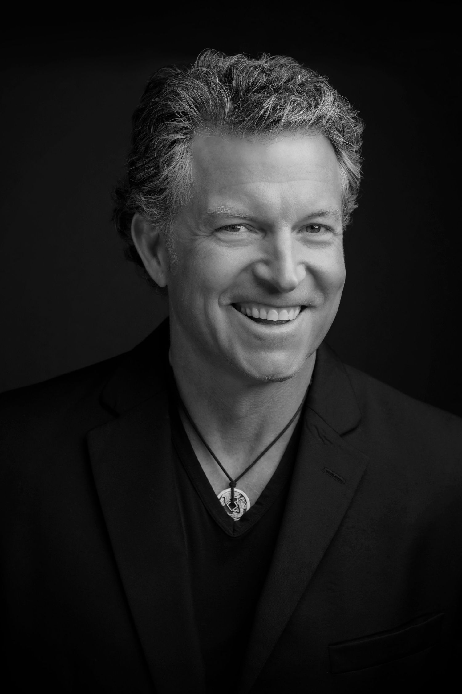 Black and white portrait of a smiling middle-aged man with gray hair wearing a blazer and pendant necklace.