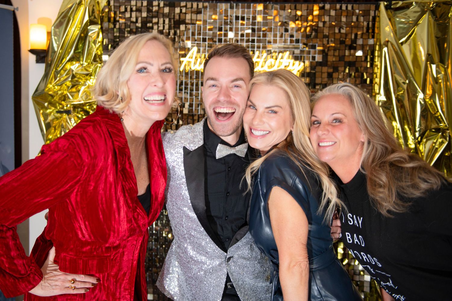 Four people smiling and posing together at a festive party with gold balloon decorations in the background.