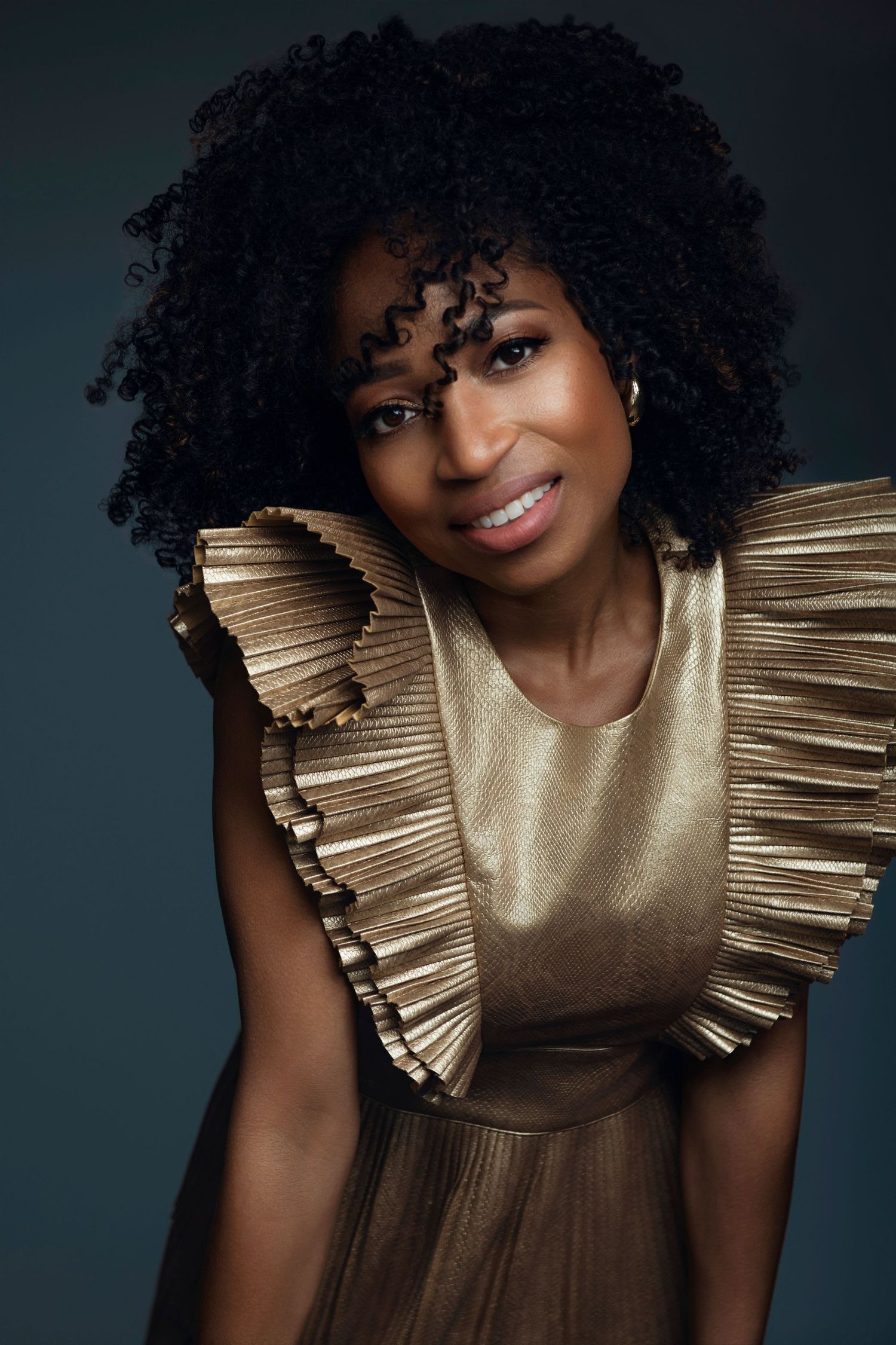 Woman with natural afro hair wearing a gold ruffled dress posing against a dark teal background.