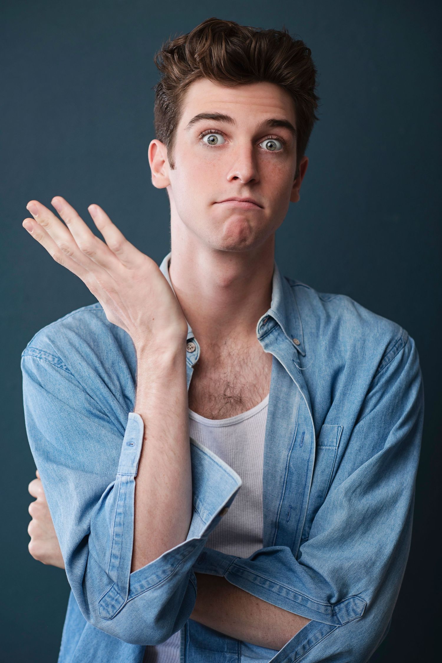 Expressive young man in a denim shirt gestures with hand raised against a dark blue background.