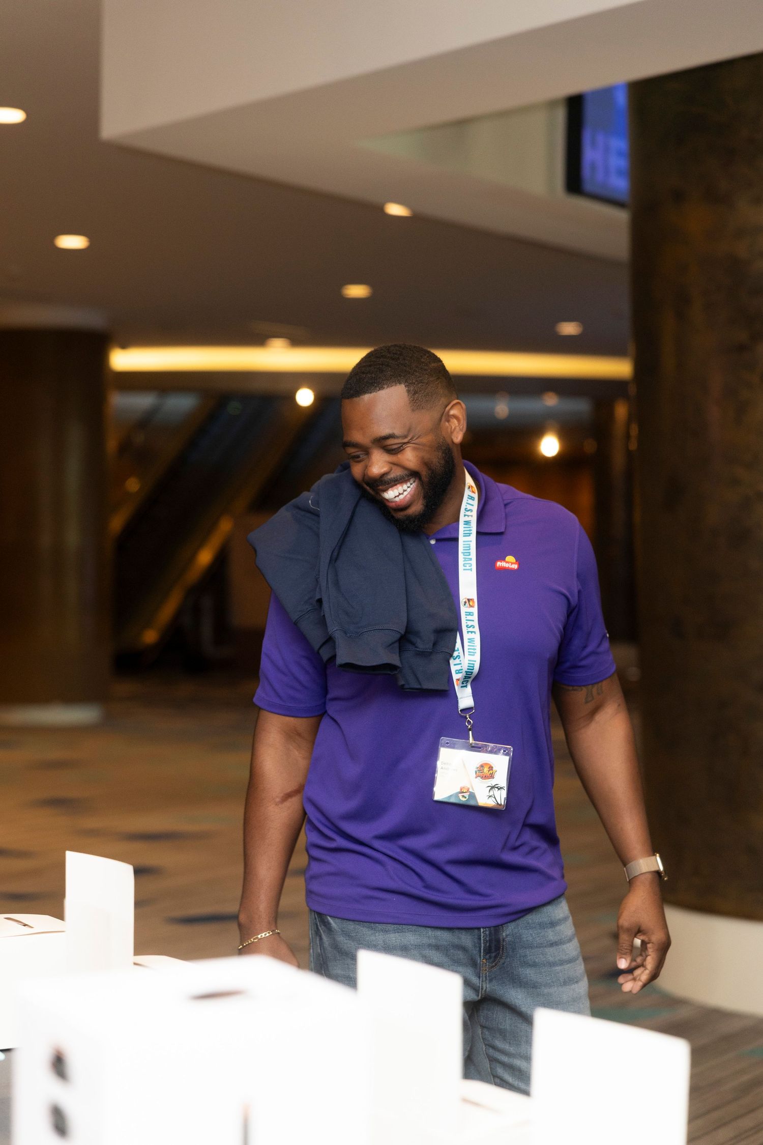 Smiling man in purple polo shirt wearing a conference lanyard badge in a modern hotel lobby.