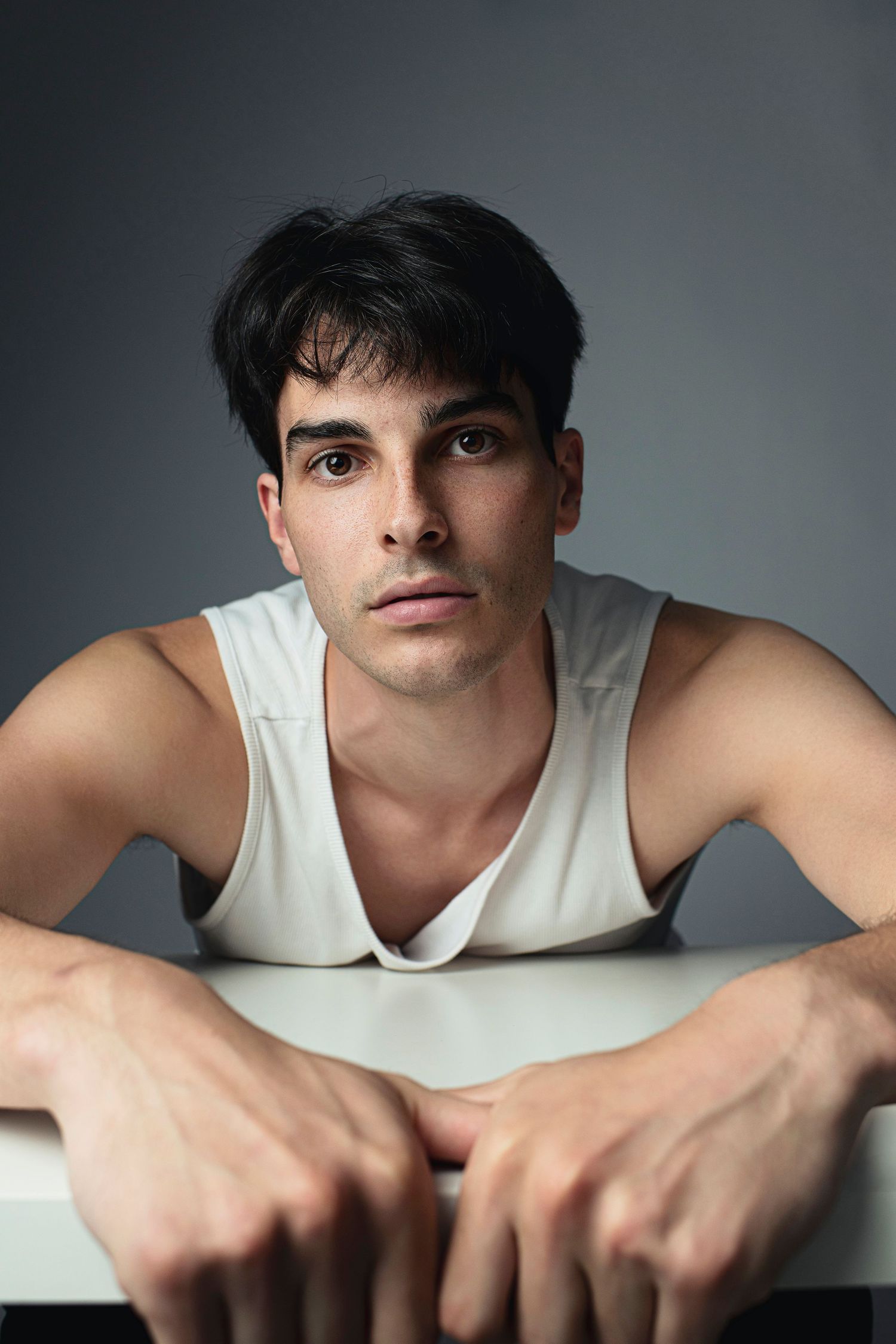 Young man in white tank top leaning on white surface with intense gaze against gray background.