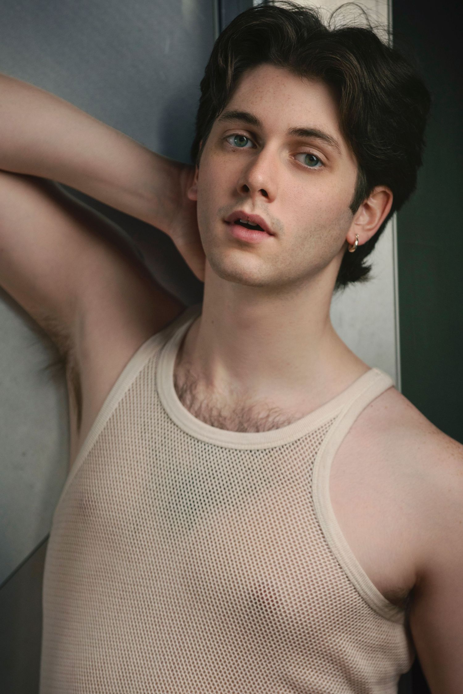 Young man in beige mesh tank top leaning against wall in artistic fashion portrait.