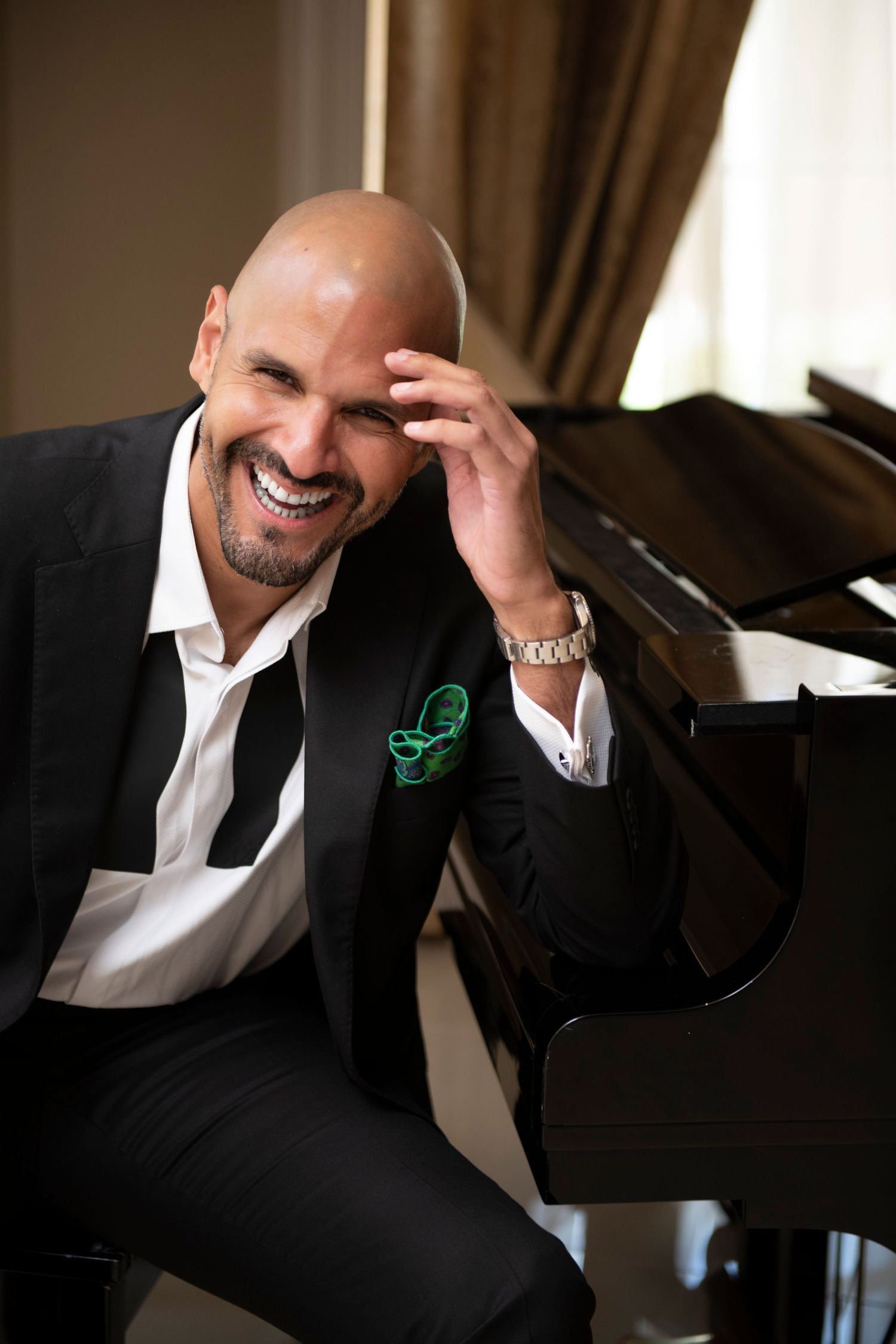 Smiling bald man in black suit with green pocket square sits at grand piano, laughing joyfully.