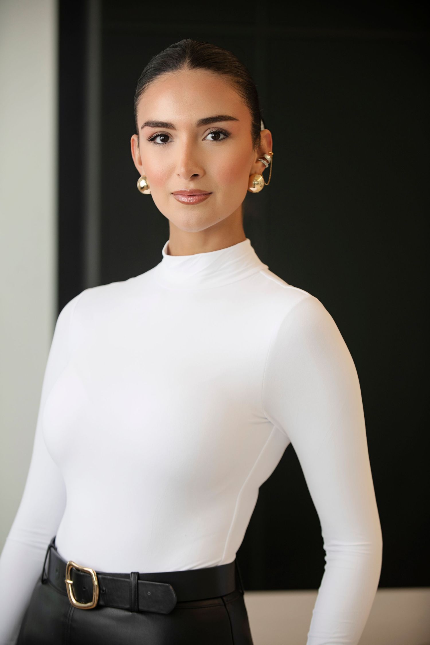 Elegant woman in white turtleneck top with gold earrings and black belt against a dark background.