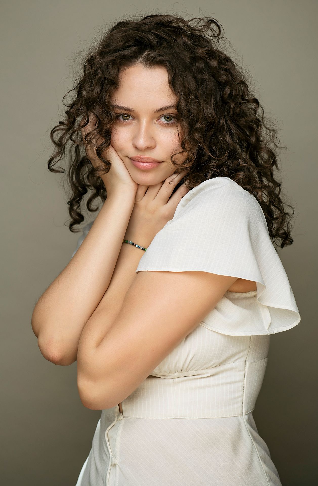 Young woman with curly brown hair posing in a white dress with hands framing her face.