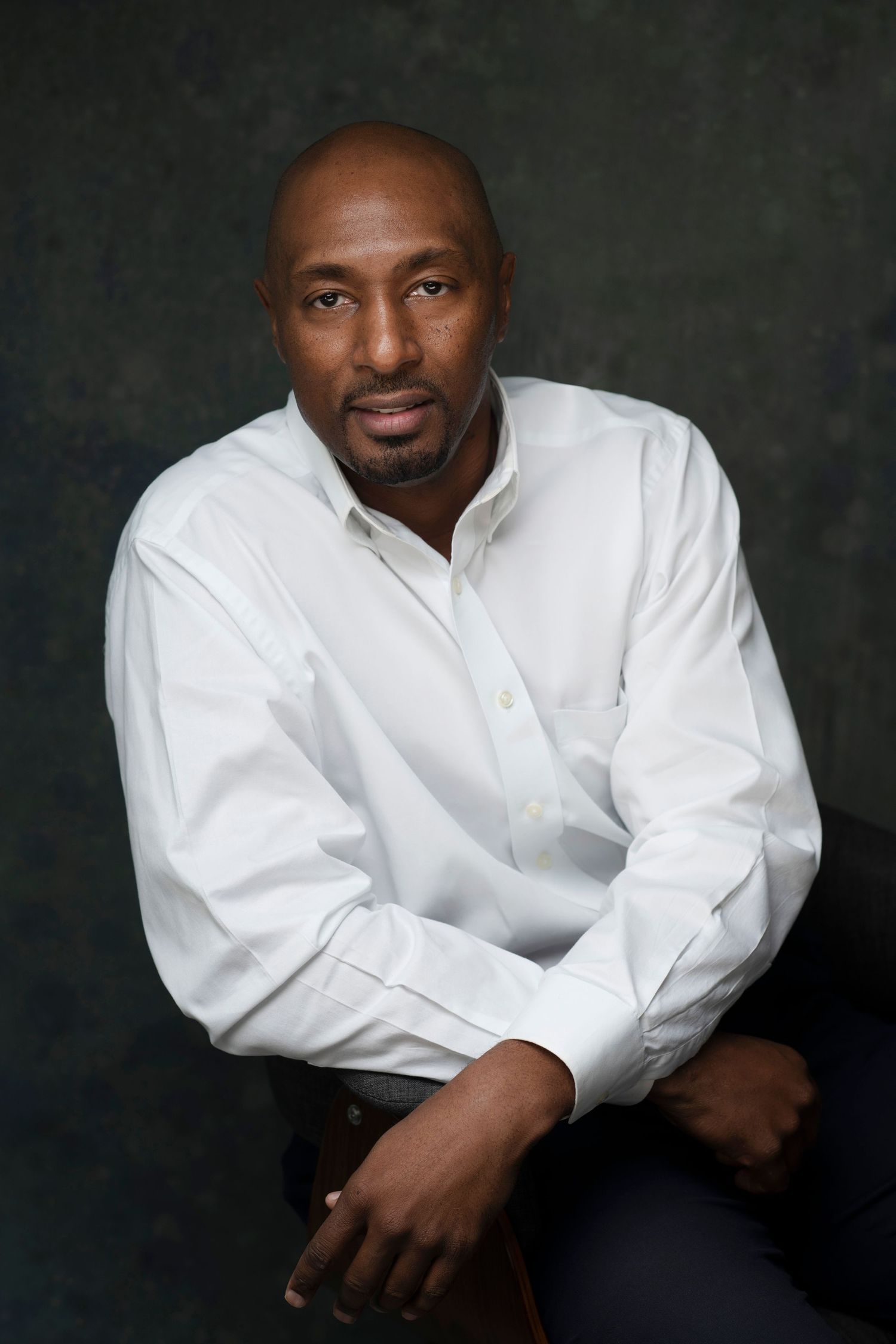 Confident bald Black man in white dress shirt seated against dark background in professional portrait.